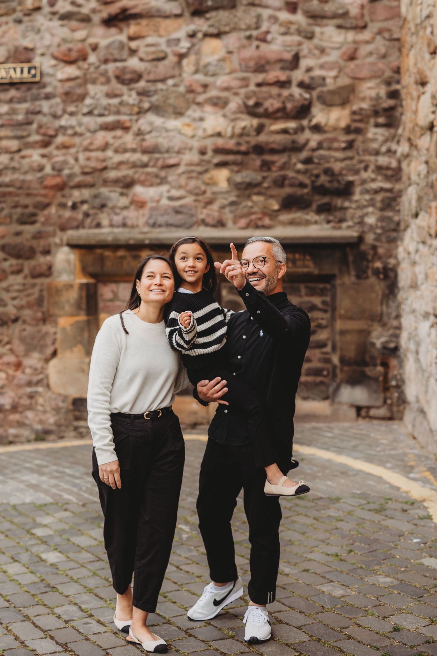 Family of three posing in front of a historic stone wall, father pointing upward while holding child.