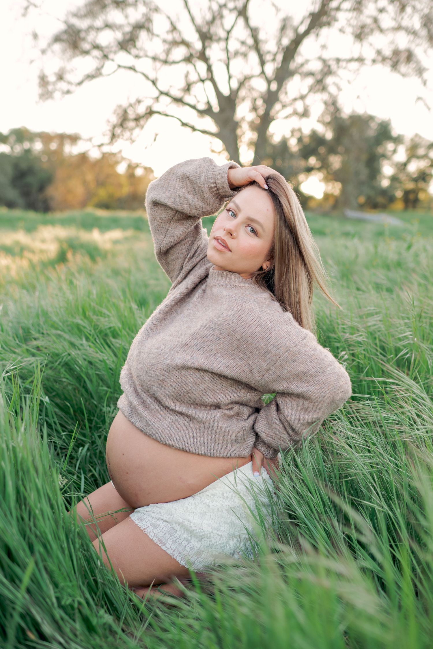 Pregnant woman in brown sweater and white shorts sitting in tall green grass field during golden hour sunset.