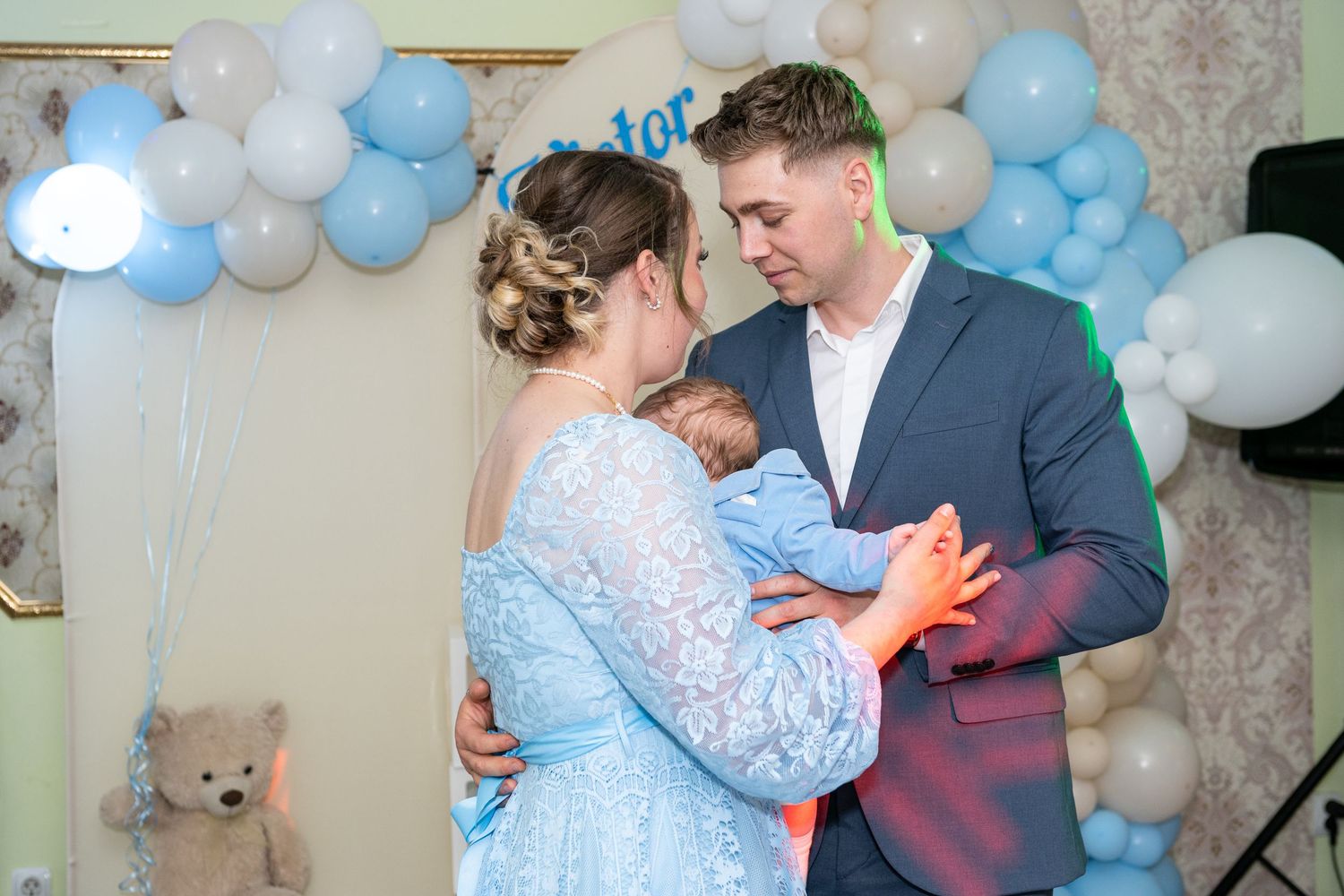 Young couple lying down with an alert baby perched between them, wearing pearl necklace and blue suit.