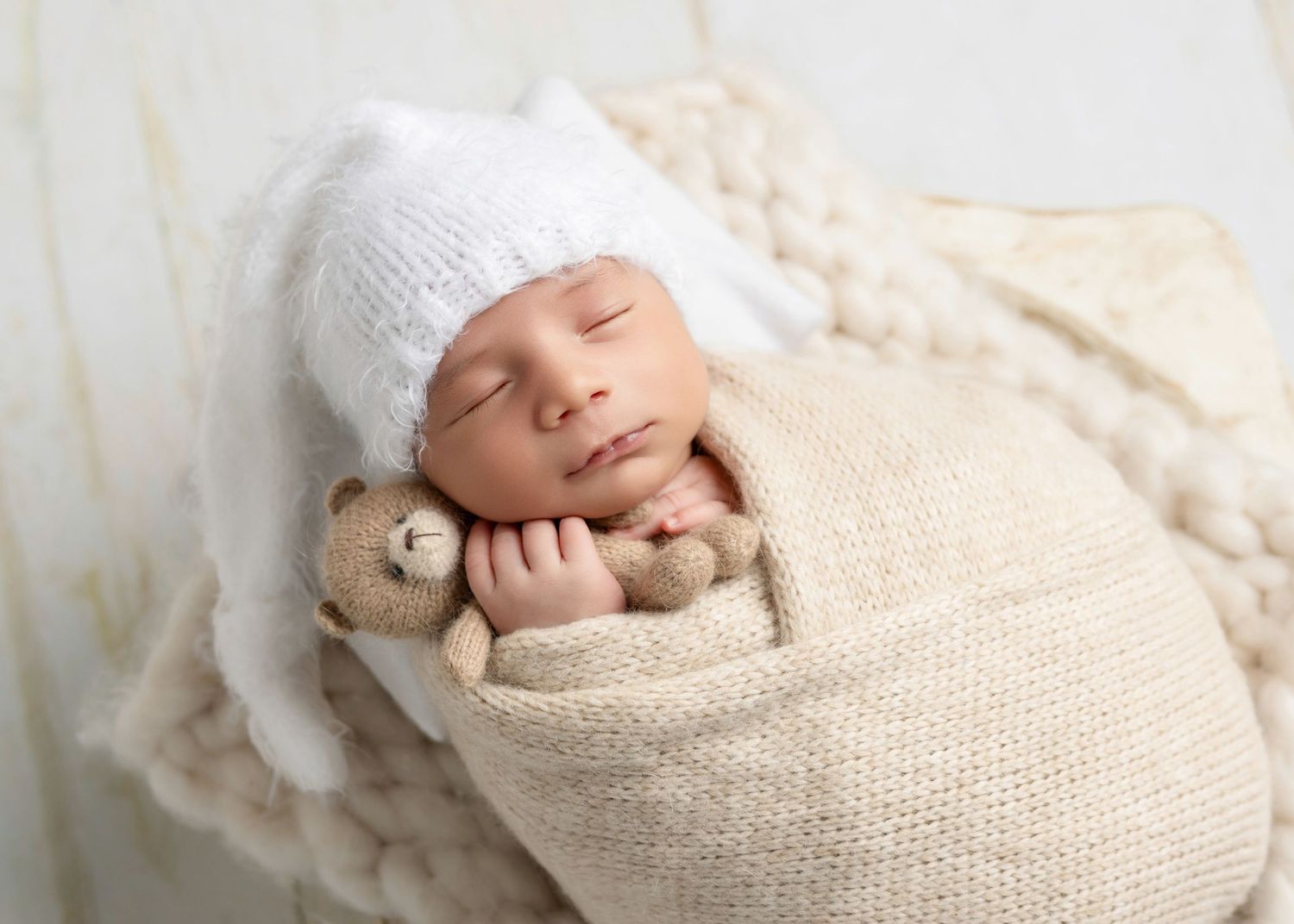 Adorable baby in brown knit overalls sitting in a wooden chair with green blanket against blue backdrop.