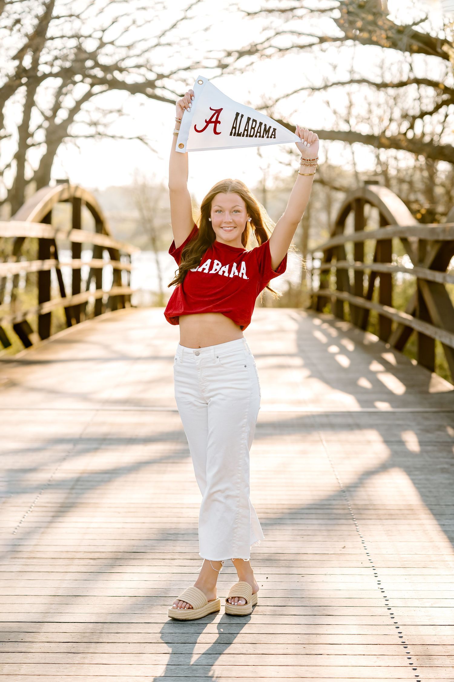 Young woman in Alabama Crimson Tide crop top holding pennant flag on wooden bridge in golden hour light.