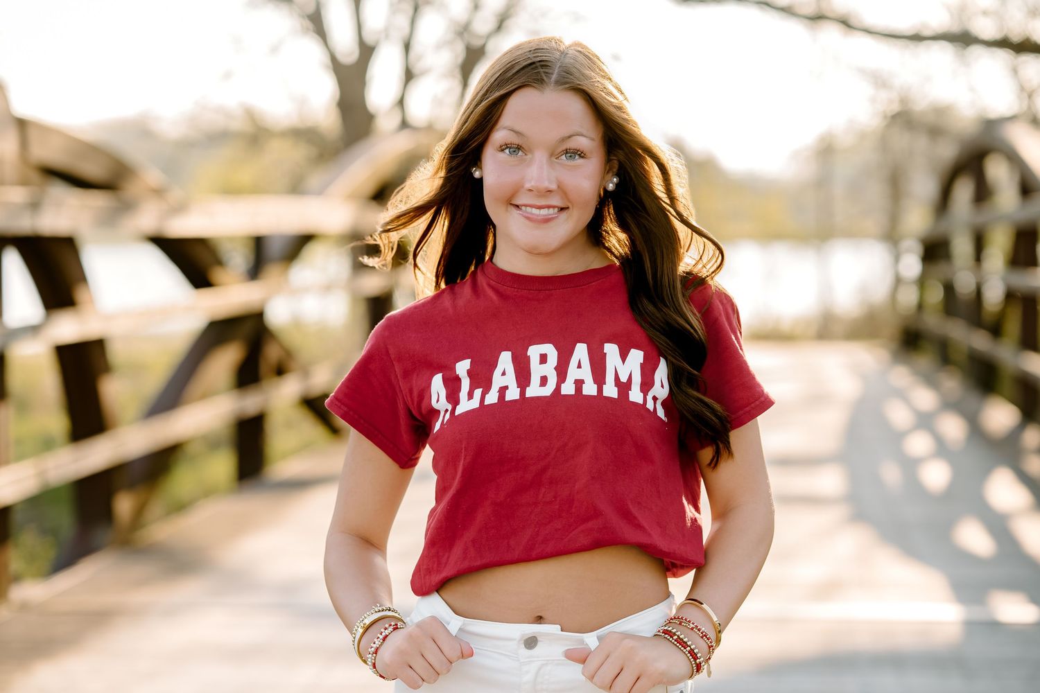 Young woman smiling in a red Alabama crop top and white shorts, standing on a bridge outdoors.