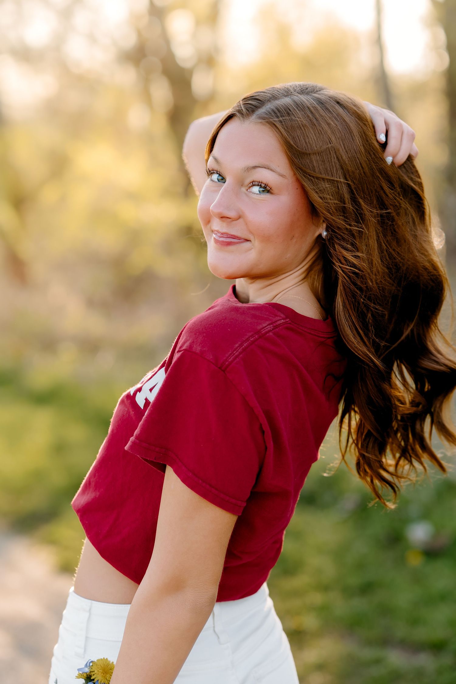 Young woman in red crop top with long brown hair poses outdoors in golden sunlight holding yellow flowers.