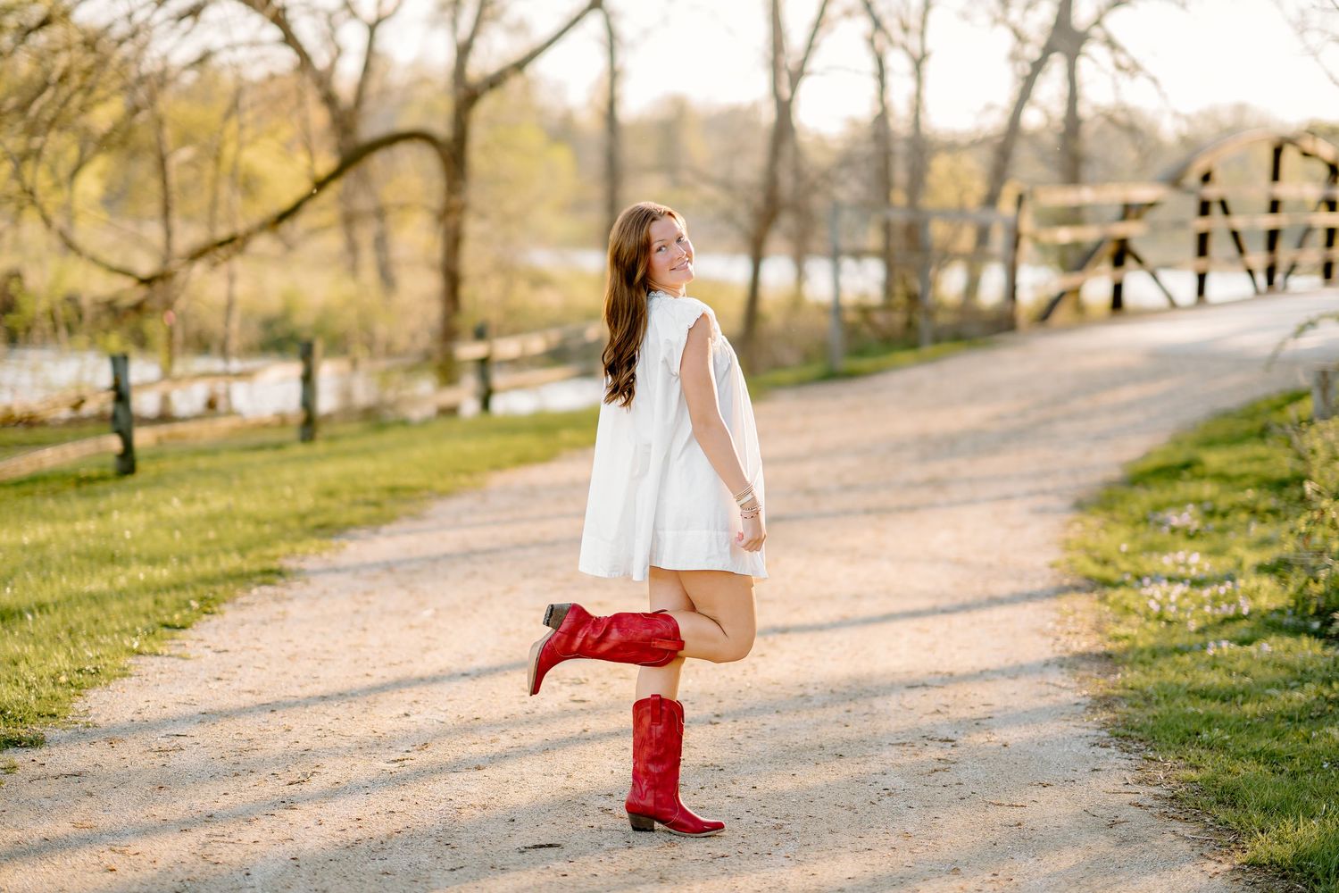 Young woman in white dress and red cowboy boots posing on a tree-lined gravel path during golden hour.