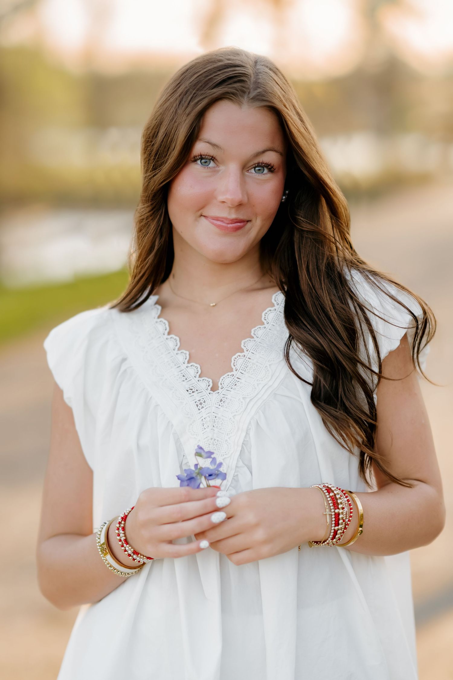 Young woman in white lace top holding purple flowers, wearing gold and red bracelets, smiling outdoors at golden hour.