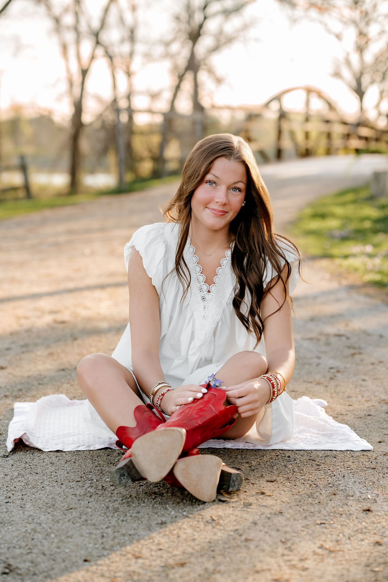 Young woman in white dress and red cowboy boots sits cross-legged on a dirt path at golden hour.