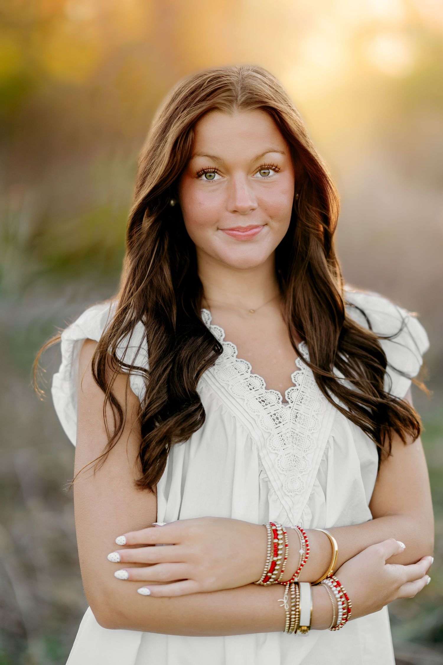 Young woman with long brown hair in white lace dress, wearing colorful bracelets, smiling outdoors at golden hour.