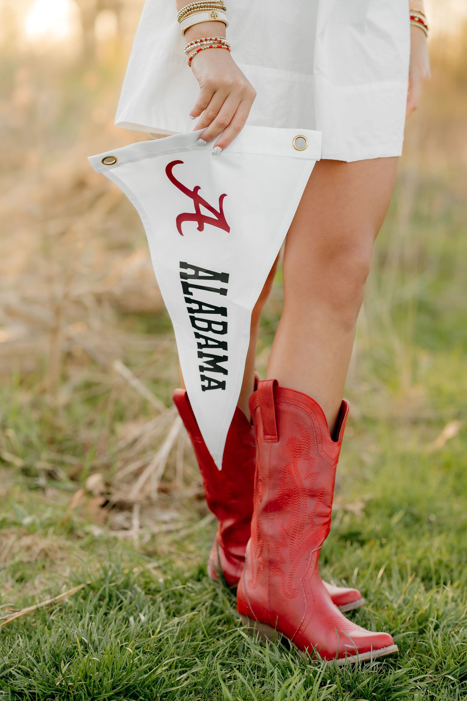 Woman in red cowboy boots holding a white Alabama Crimson Tide pennant flag while wearing a white dress outdoors.