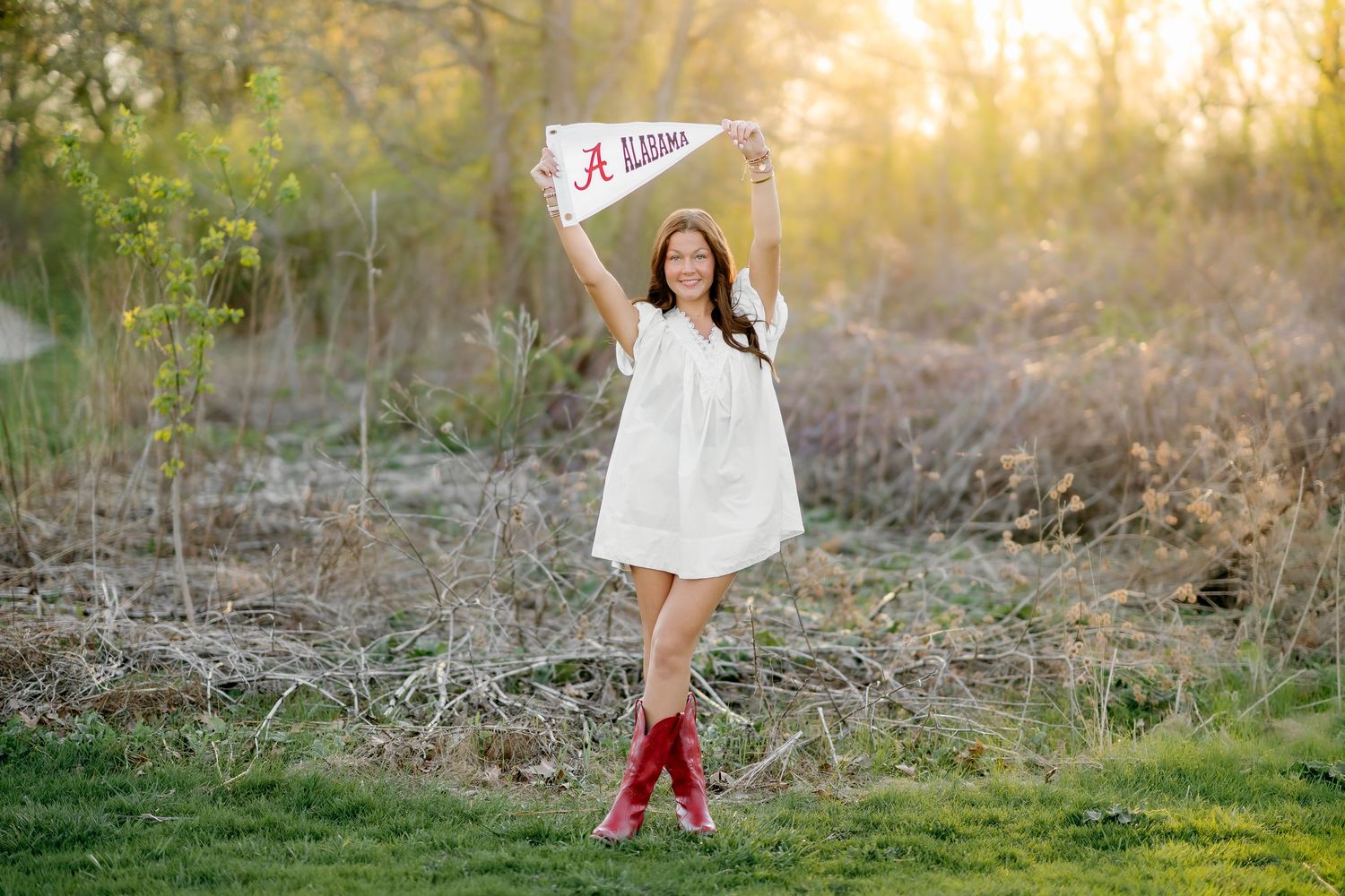 Young woman in white dress and red boots holds University of Alabama pennant flag outdoors in golden hour light.