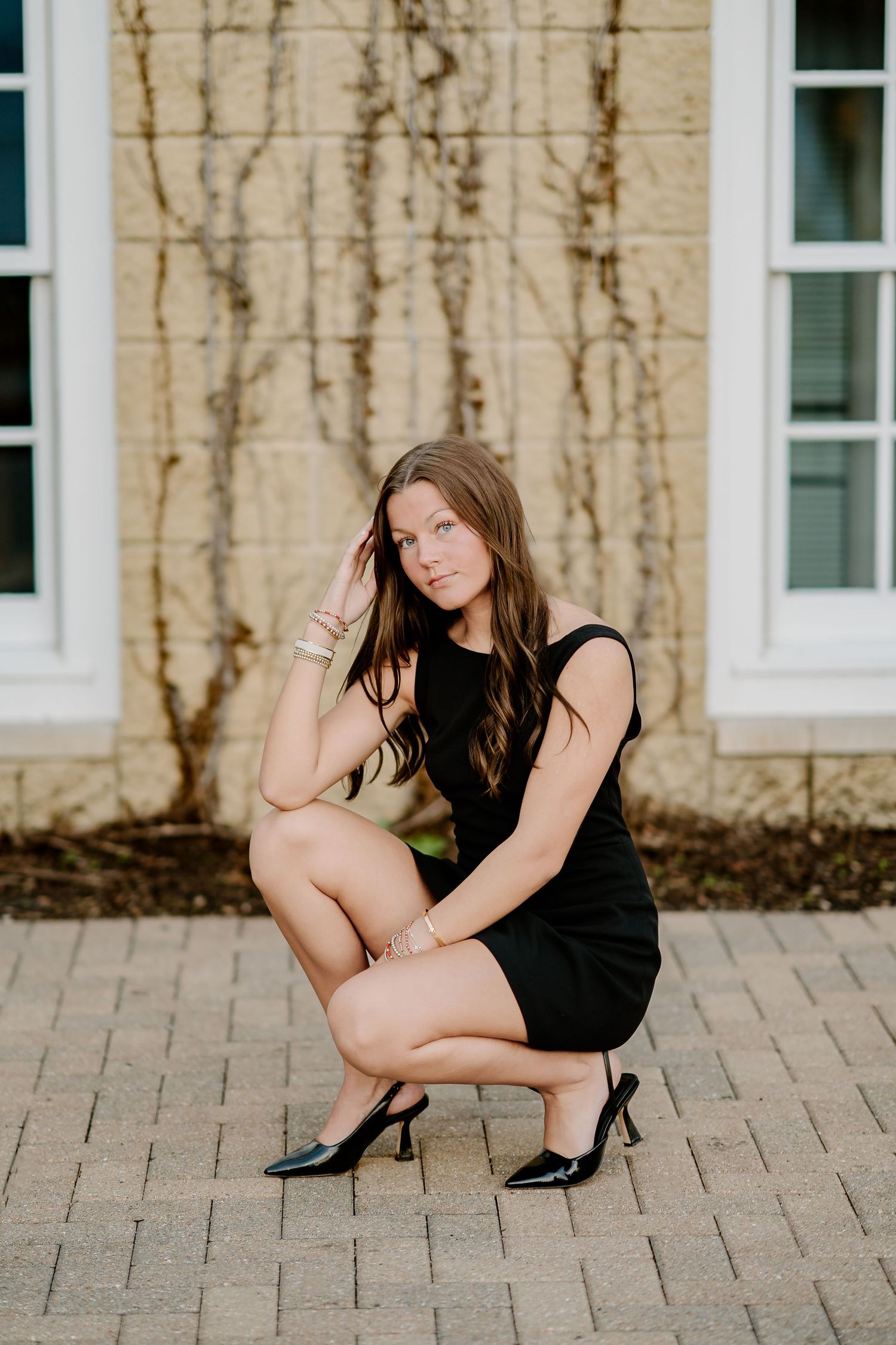 Woman in black mini dress and heels crouching on brick pavement outdoors near a building with windows.