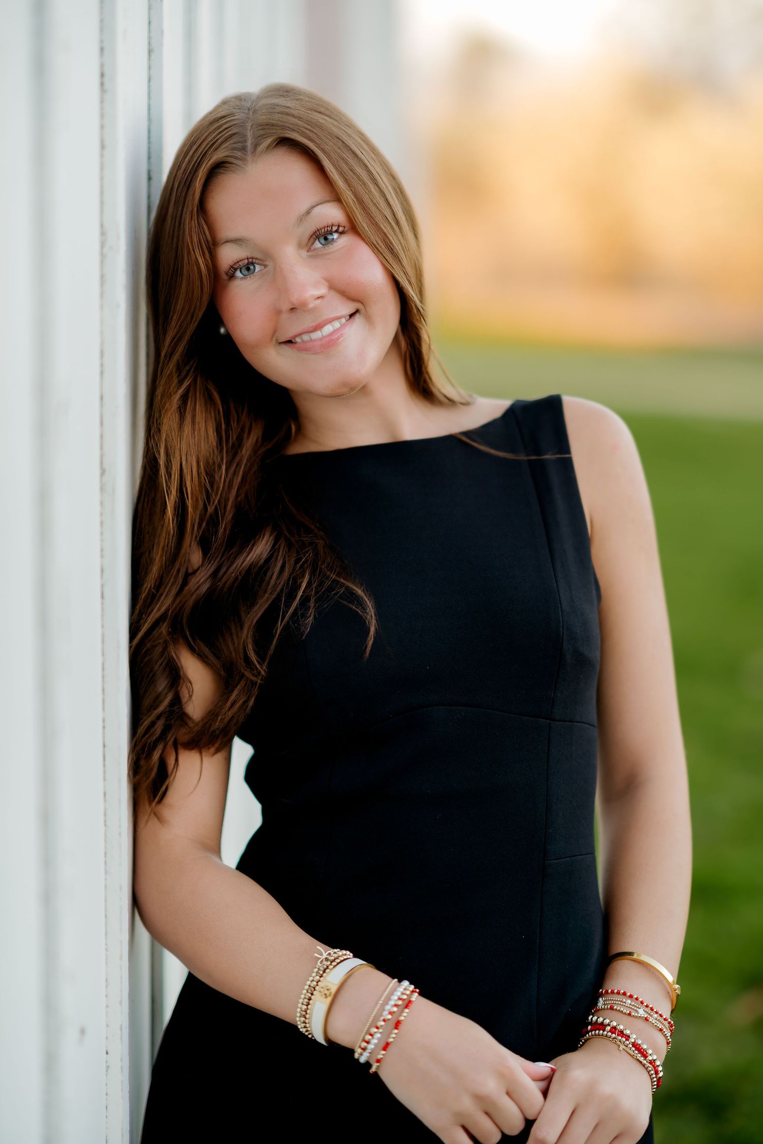 Young woman in black dress with gold bracelets smiling outdoors, leaning against white wall with green background.