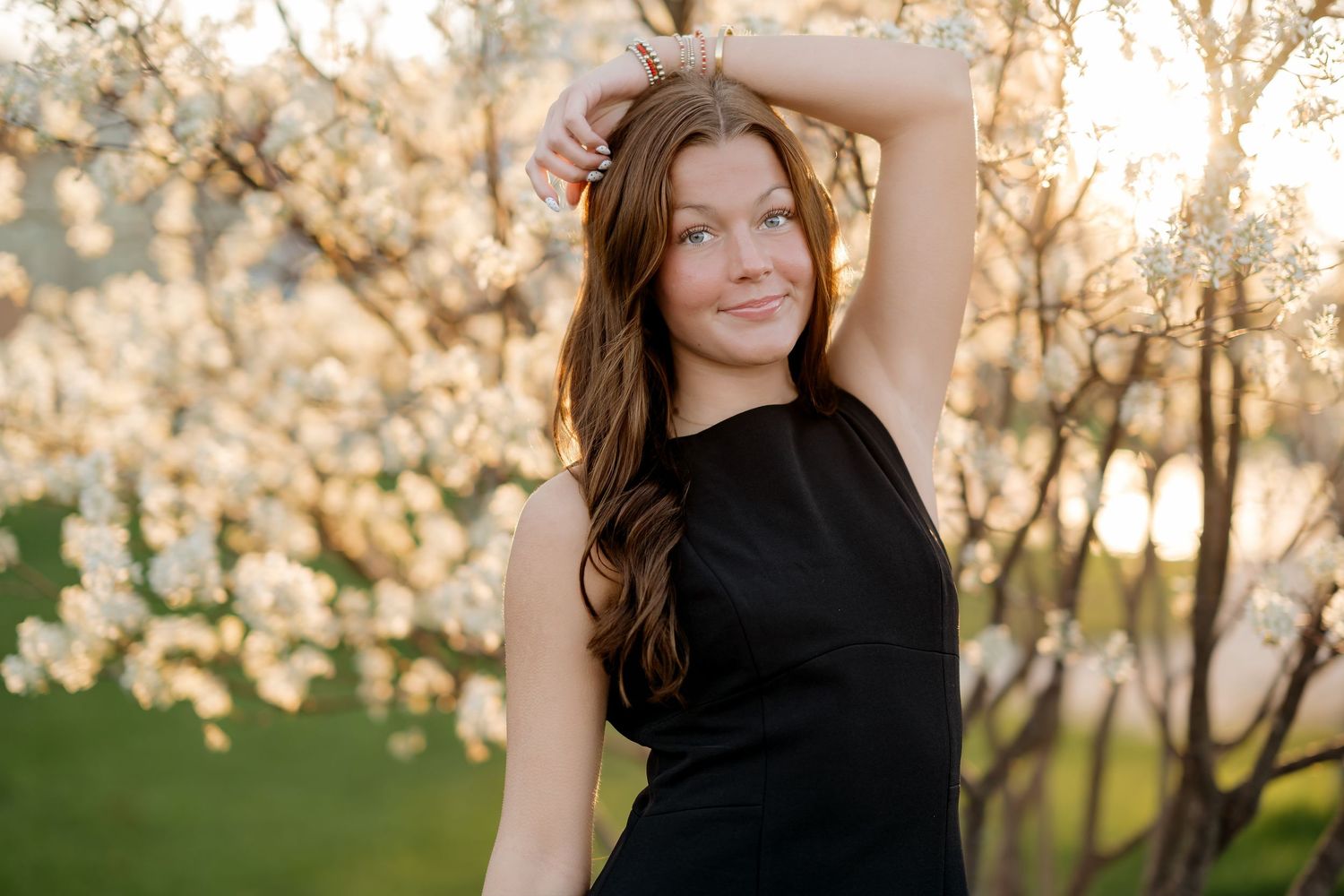 Woman in black dress posing among white flowering trees at sunset, smiling with hand raised above head.