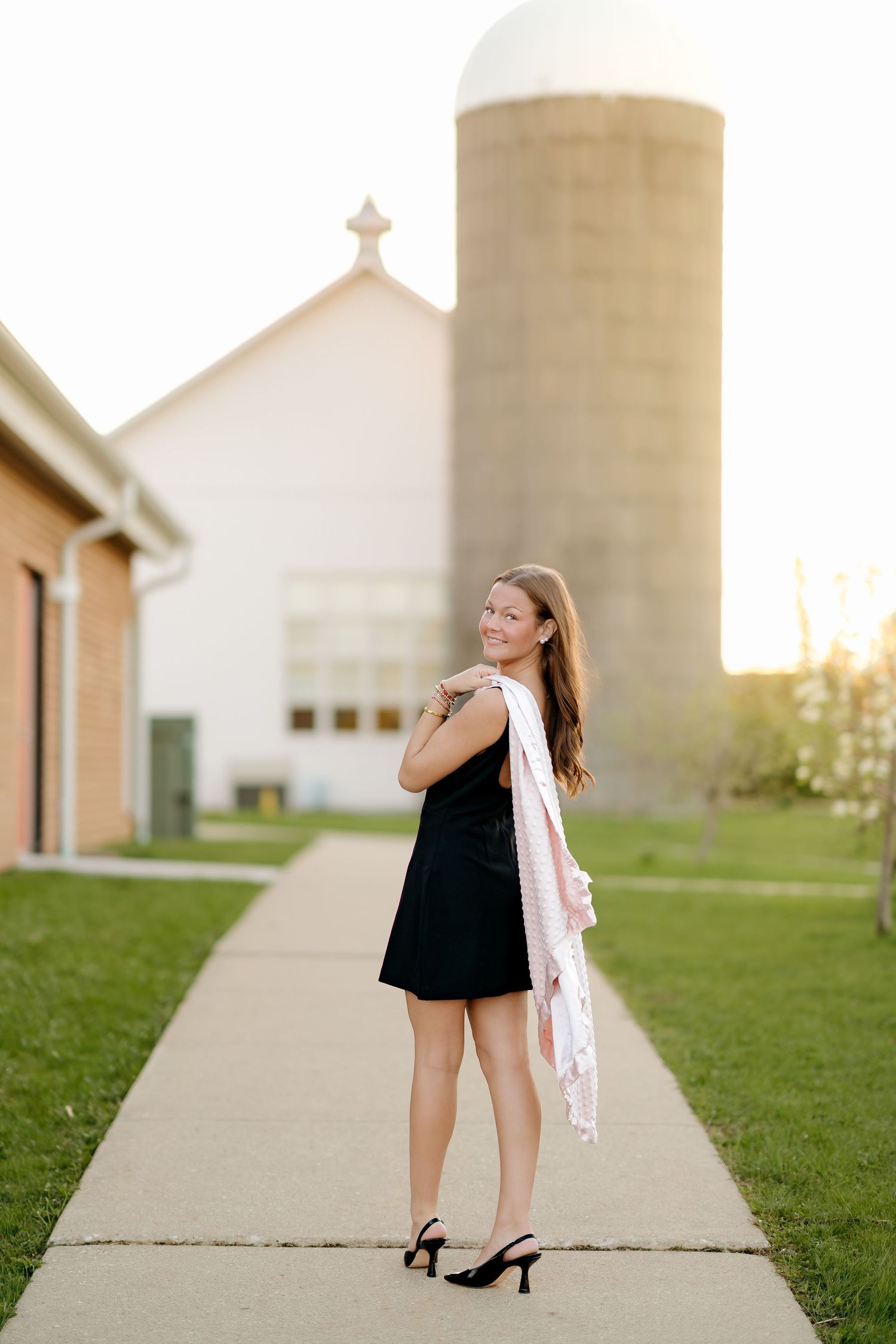Young woman in black dress with pink scarf posing on sidewalk near white barn and grain silo at sunset.