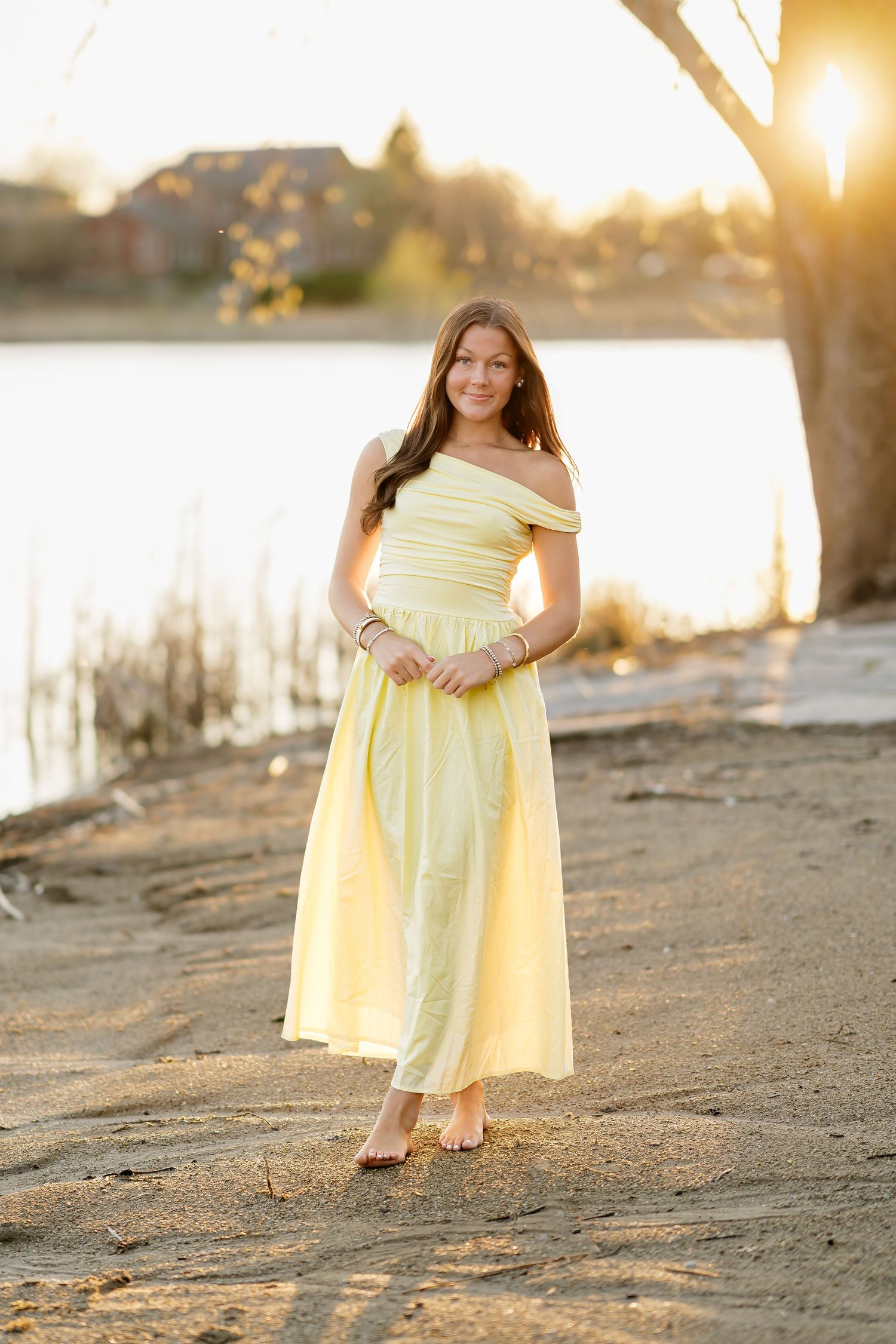 Young woman in yellow off-shoulder maxi dress standing on sandy beach at golden hour sunset.