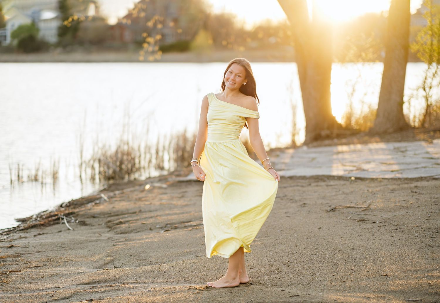 Young woman in flowing yellow dress dancing barefoot on sandy beach at golden sunset near calm water.