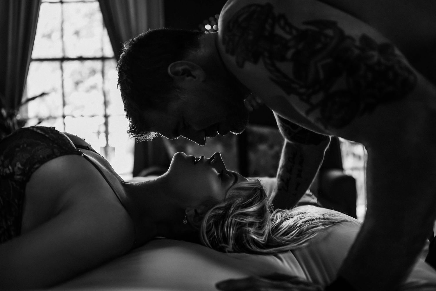 Black and white intimate couple portrait, tattooed man leaning over woman in romantic close embrace indoors. relationship boudoir session