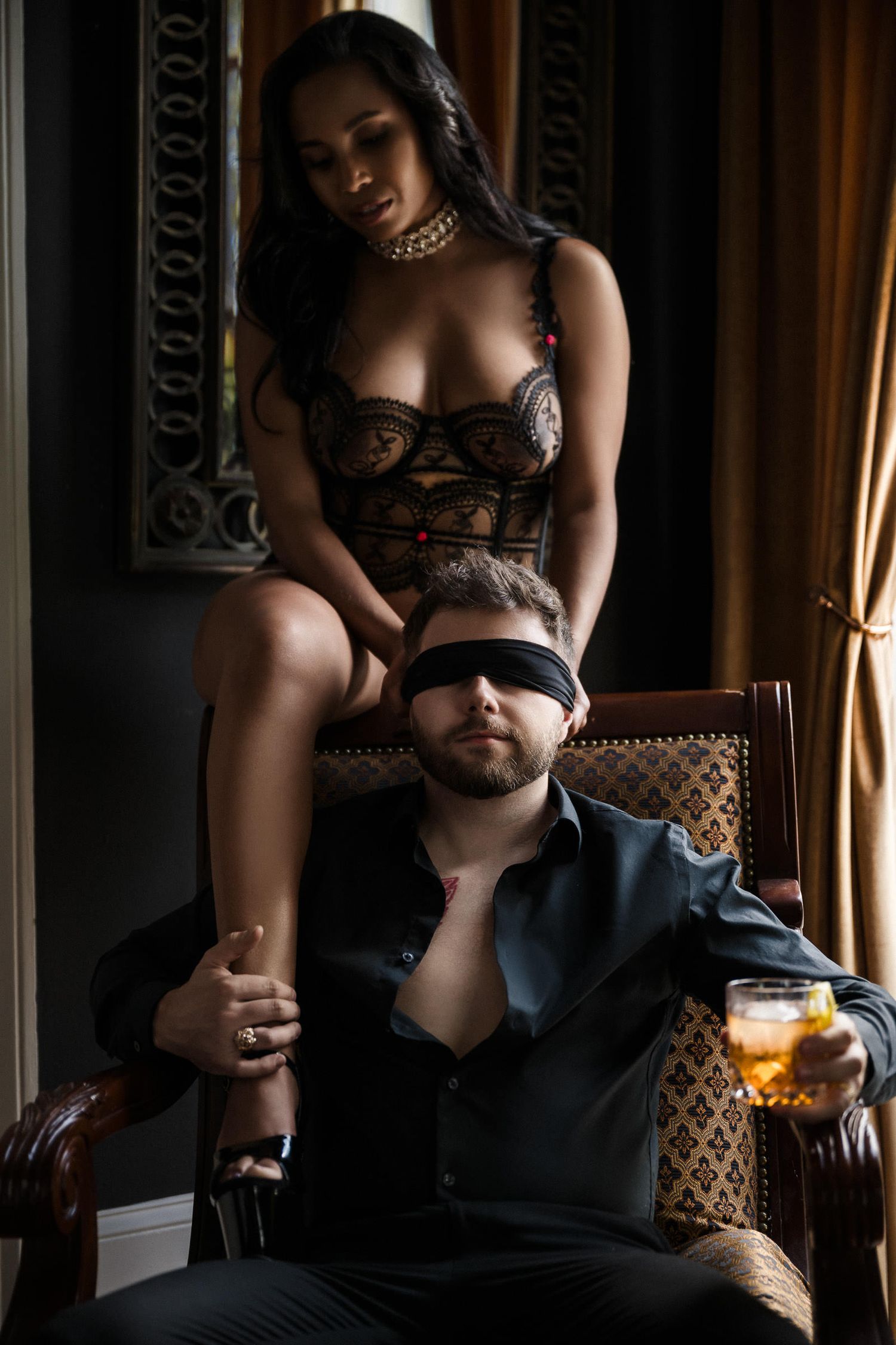 A blindfolded man in a chair while a woman in lingerie poses behind him in a moody interior. intimate couples photoshoot New Orleans.