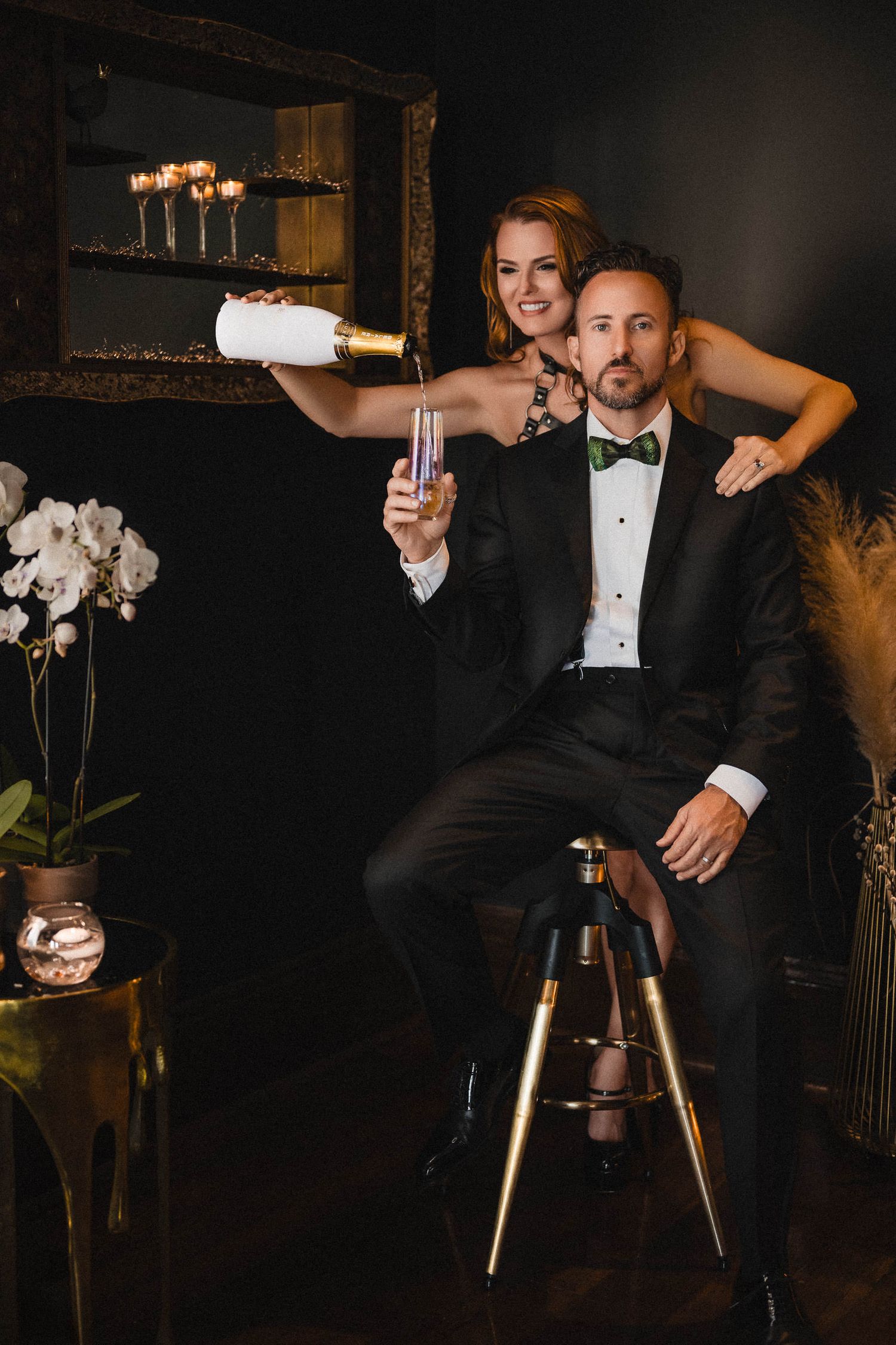 Elegant couple in formal attire celebrating with champagne, man in tuxedo seated while woman stands behind him. sensual couples photography