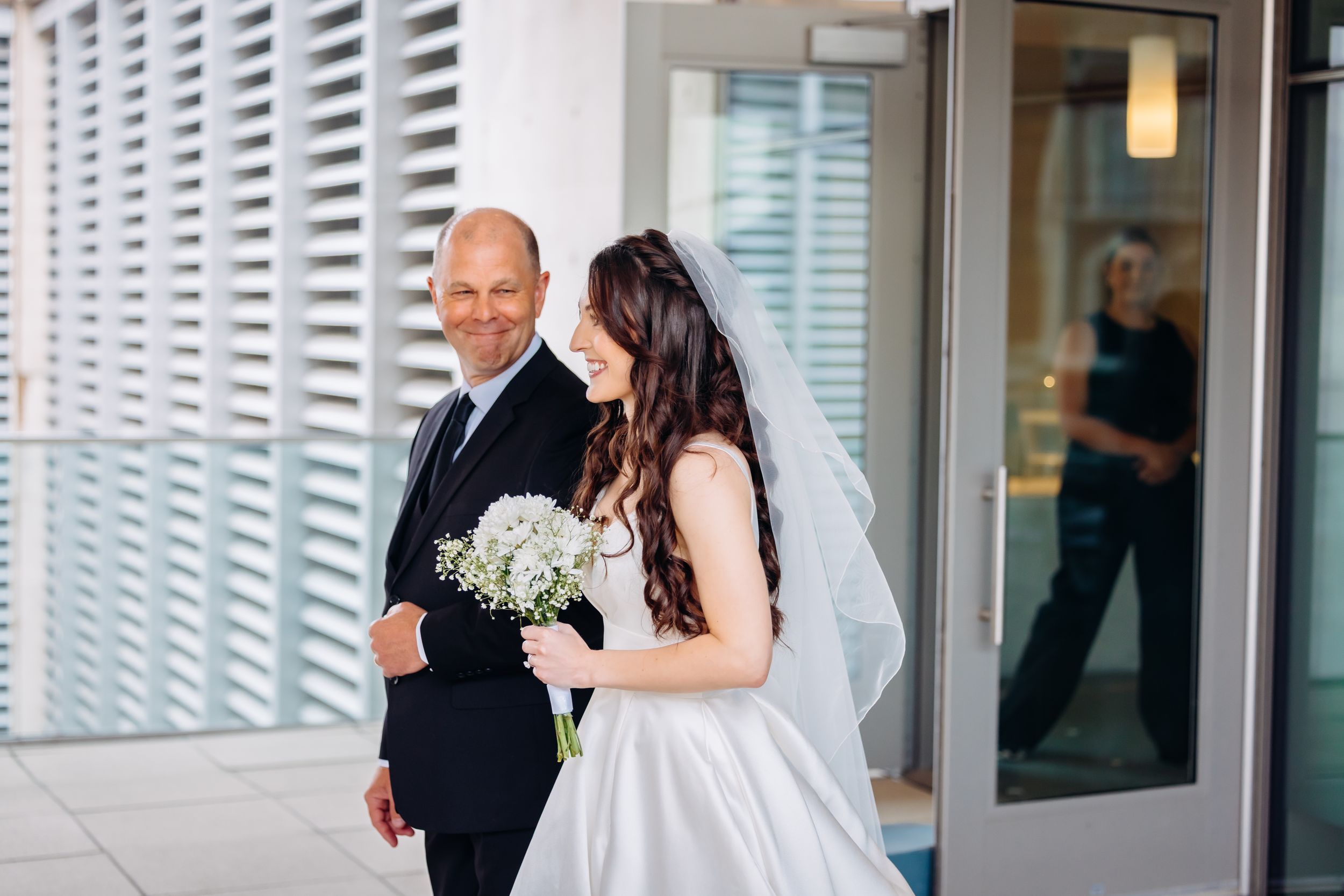 Bride in white gown holding bouquet walks with smiling man in dark suit outside modern building.