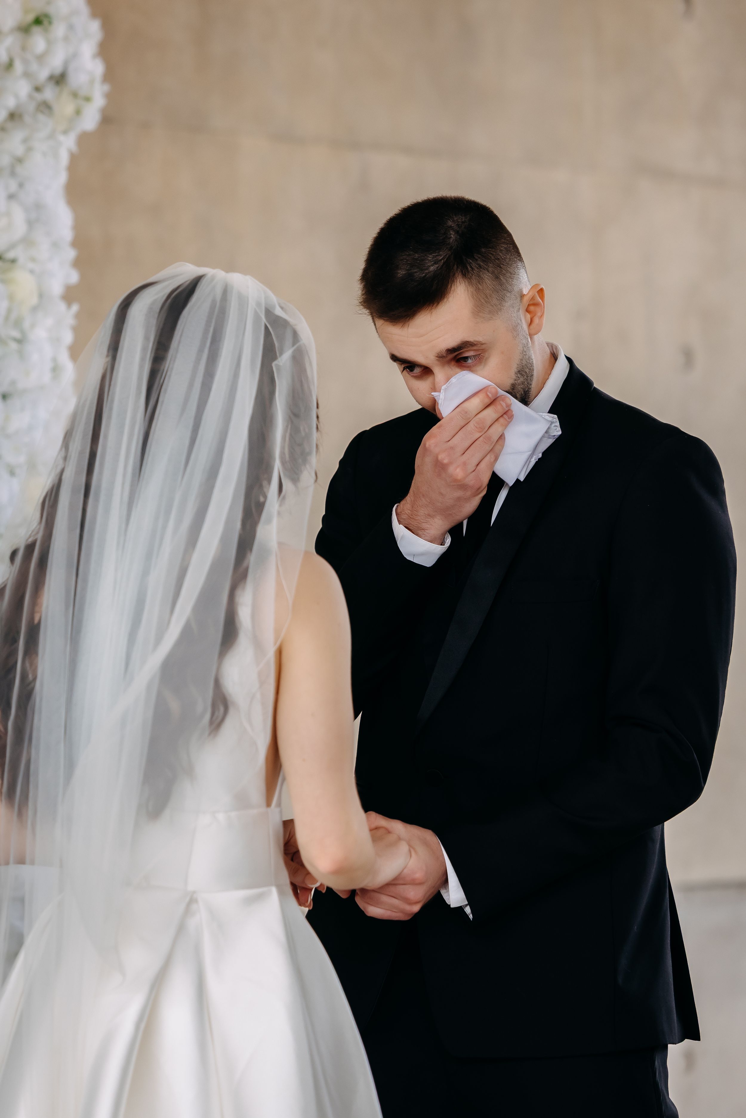 Emotional groom wiping tears with handkerchief while holding brides hands during wedding ceremony.
