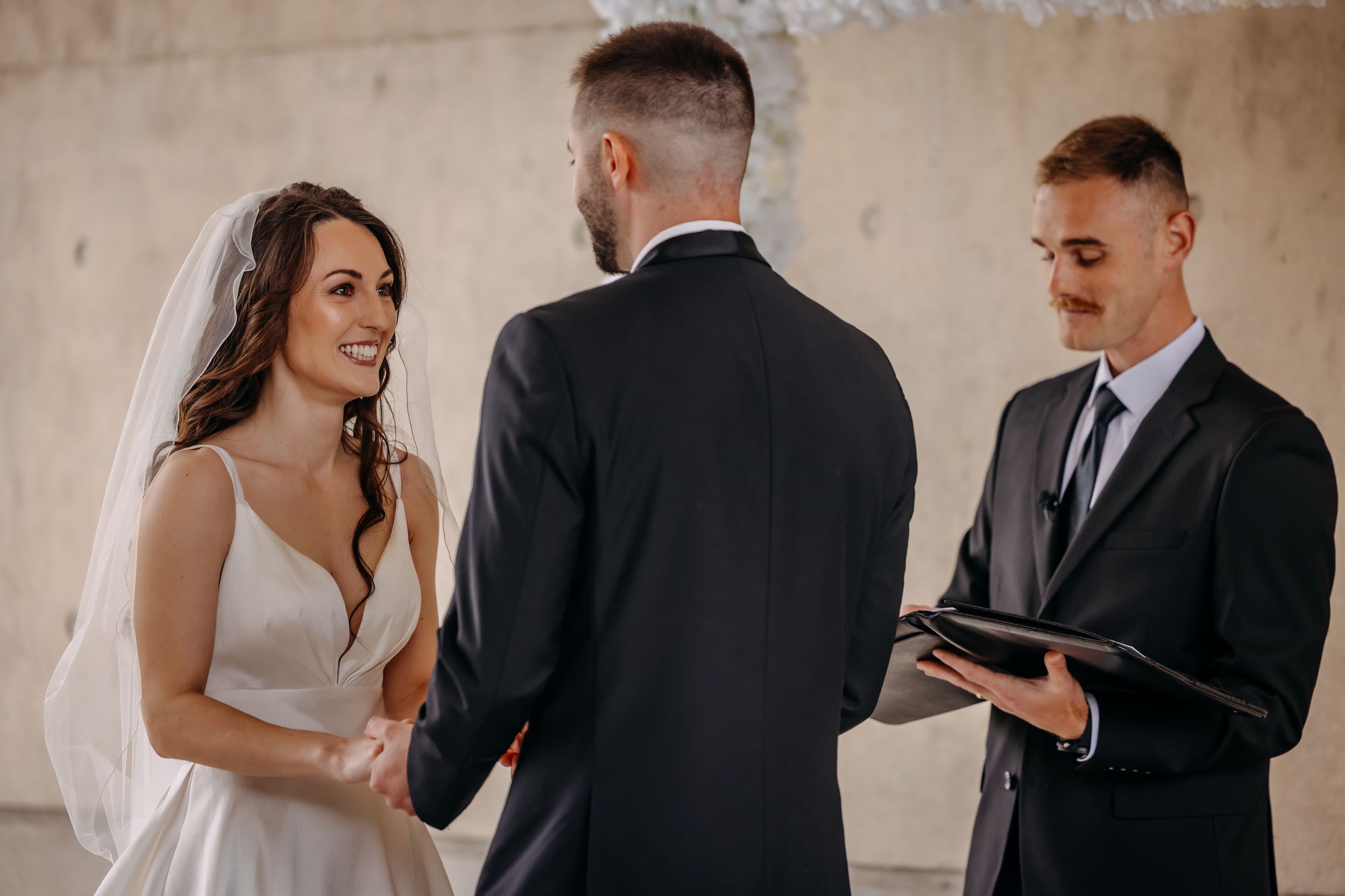 Bride and groom exchanging vows at wedding ceremony with officiant holding book indoors.