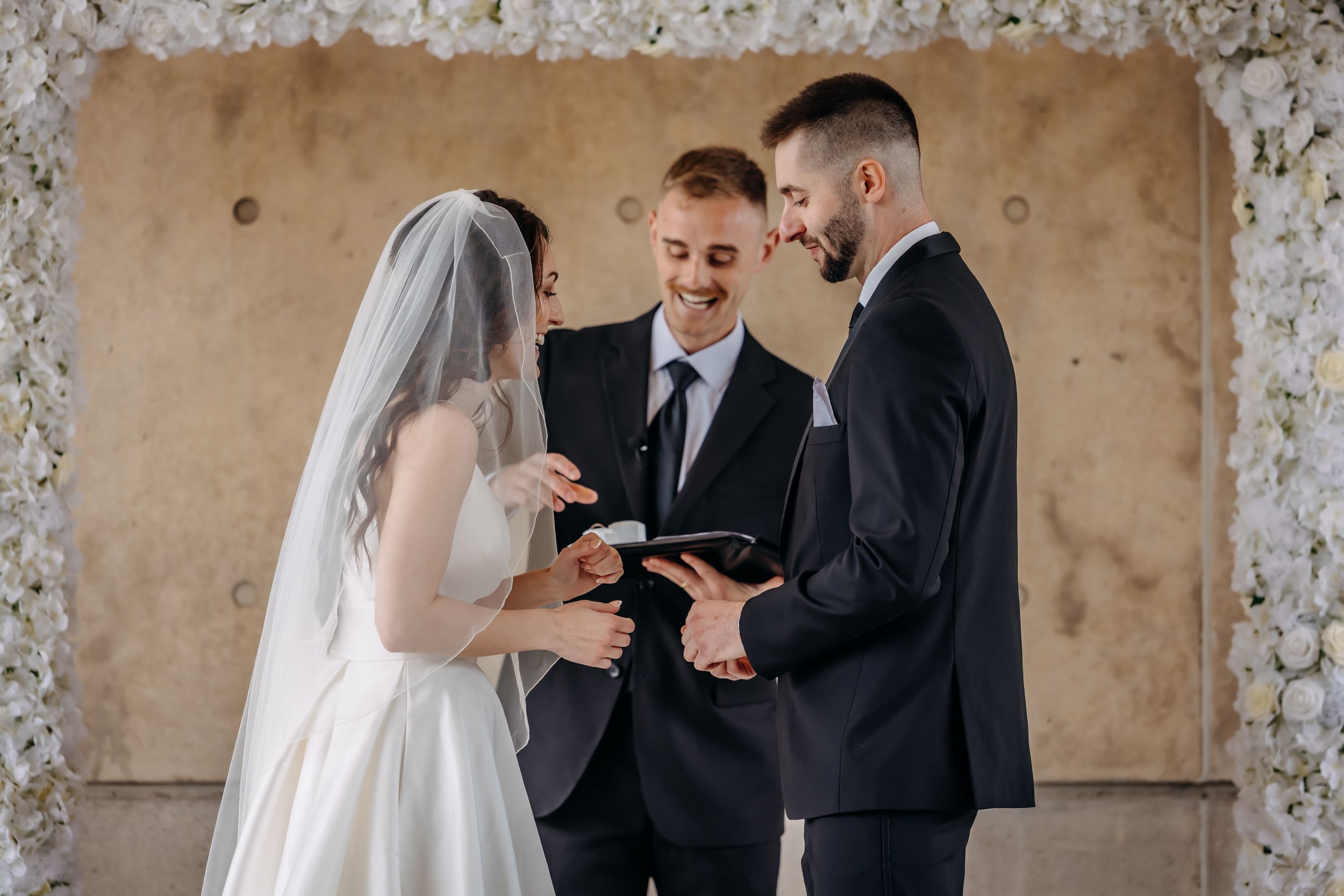 A bride and groom exchange rings during a wedding ceremony with a smiling officiant holding a book before a floral arch.