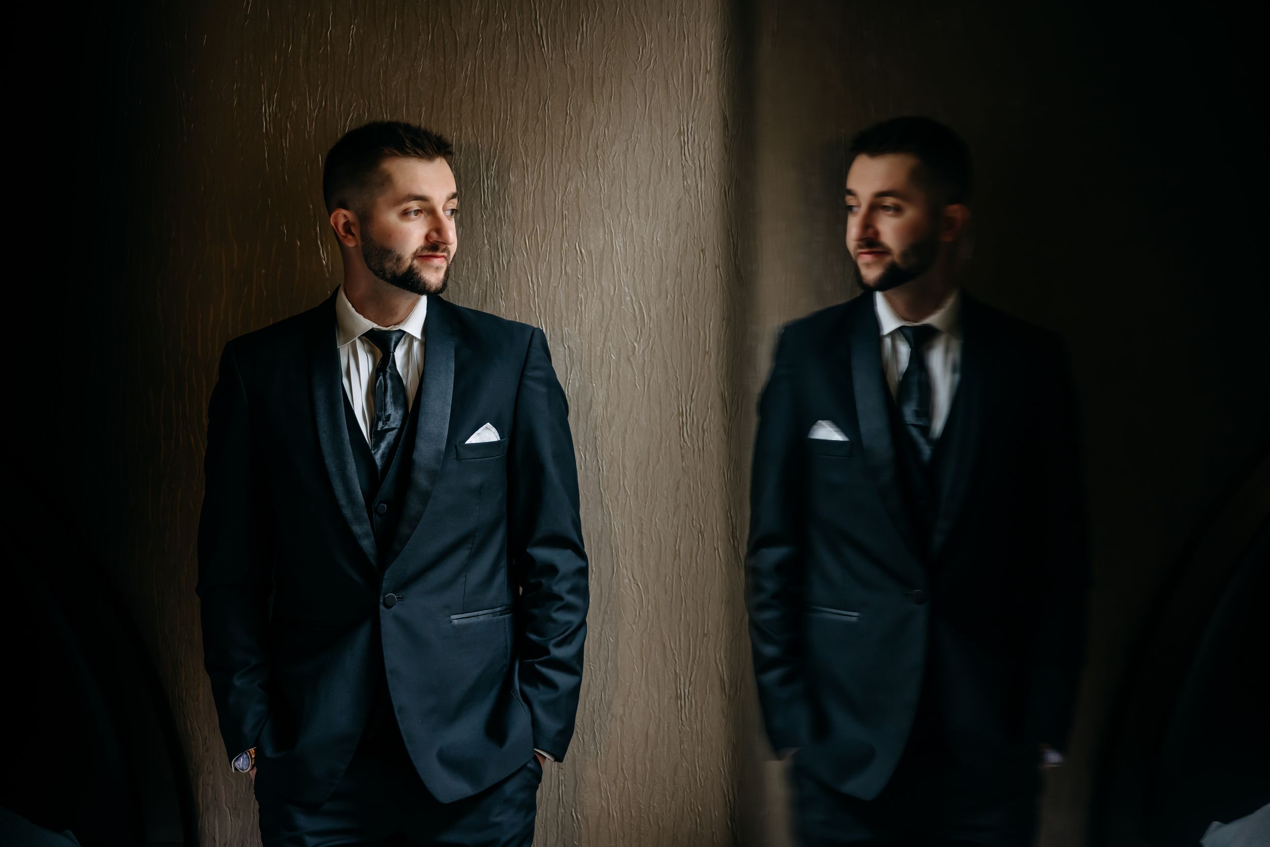 A groom in a dark suit with white pocket square stands beside his mirror reflection against a wooden wall.