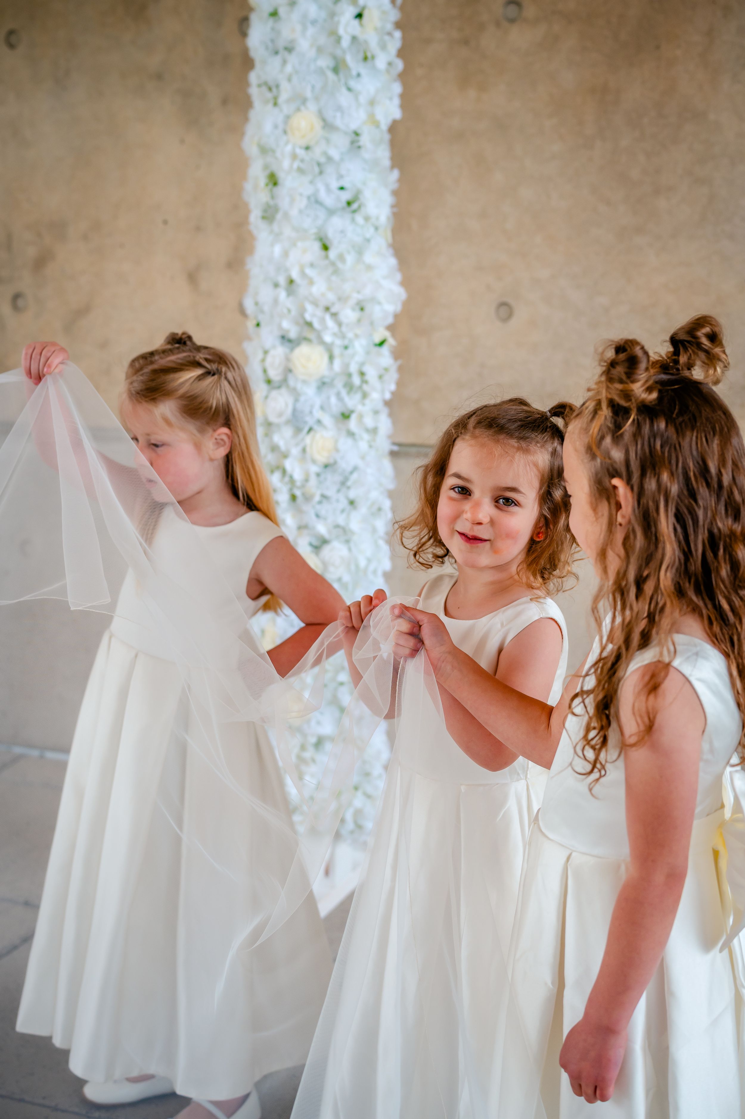 Three young flower girls in white dresses hold a bridal veil near a floral column at a wedding ceremony.