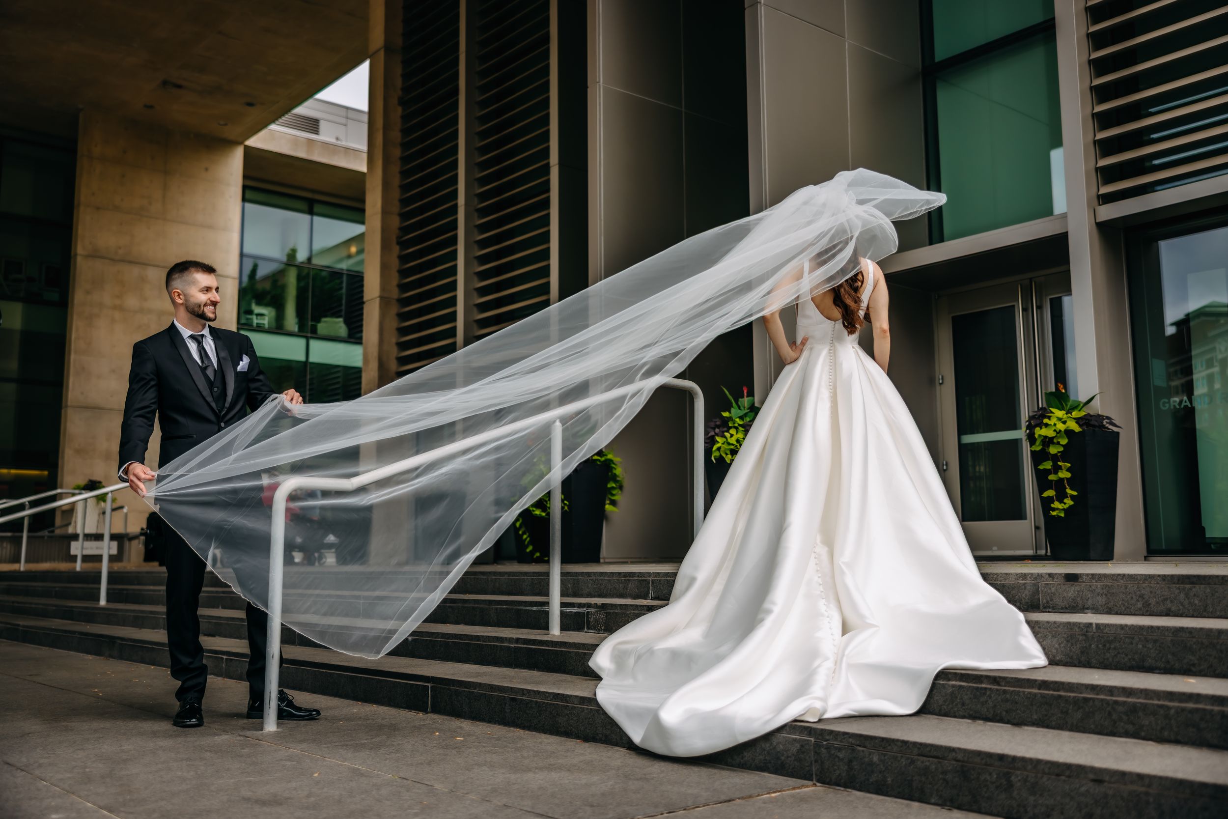 Groom holds bride's flowing veil on wedding day outside modern building with steps.