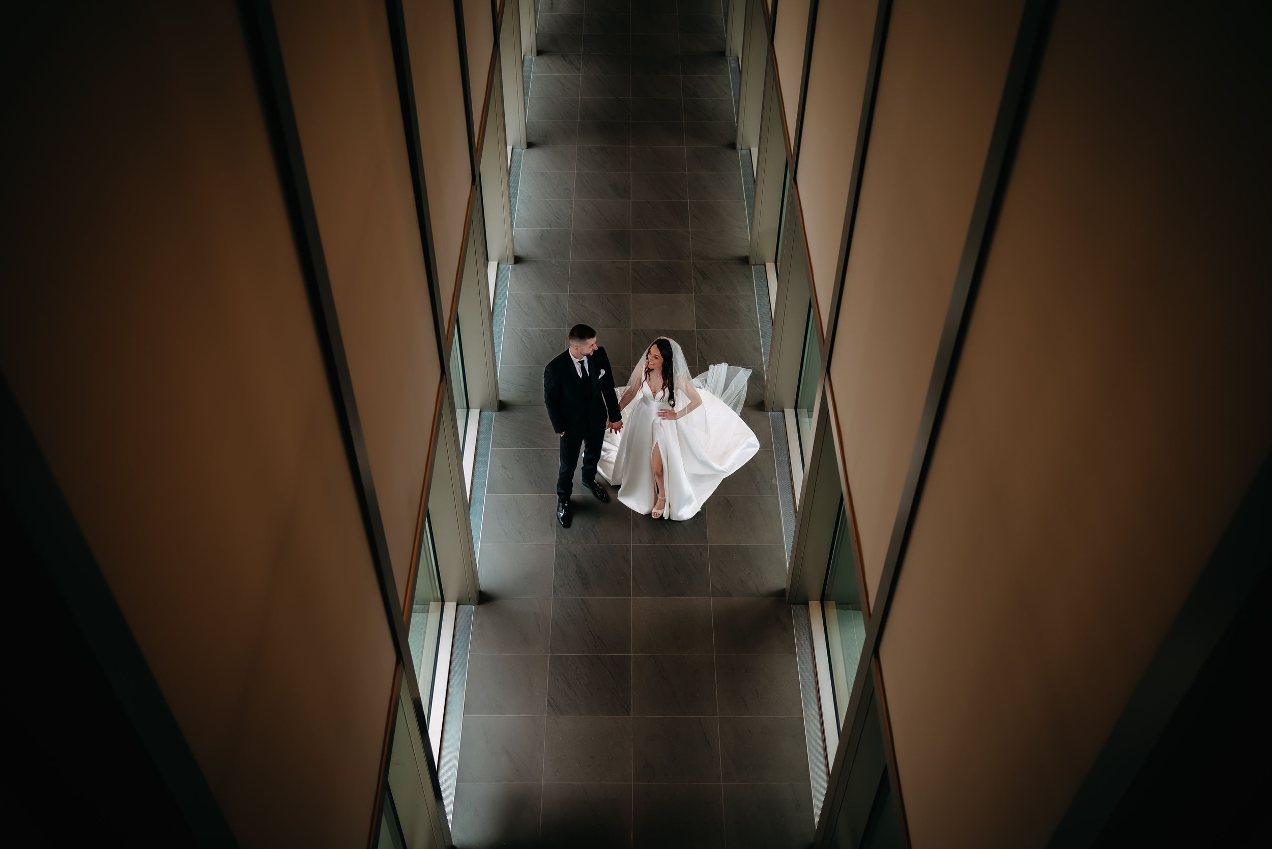 Aerial view of bride and groom walking through modern architectural corridor on wedding day.