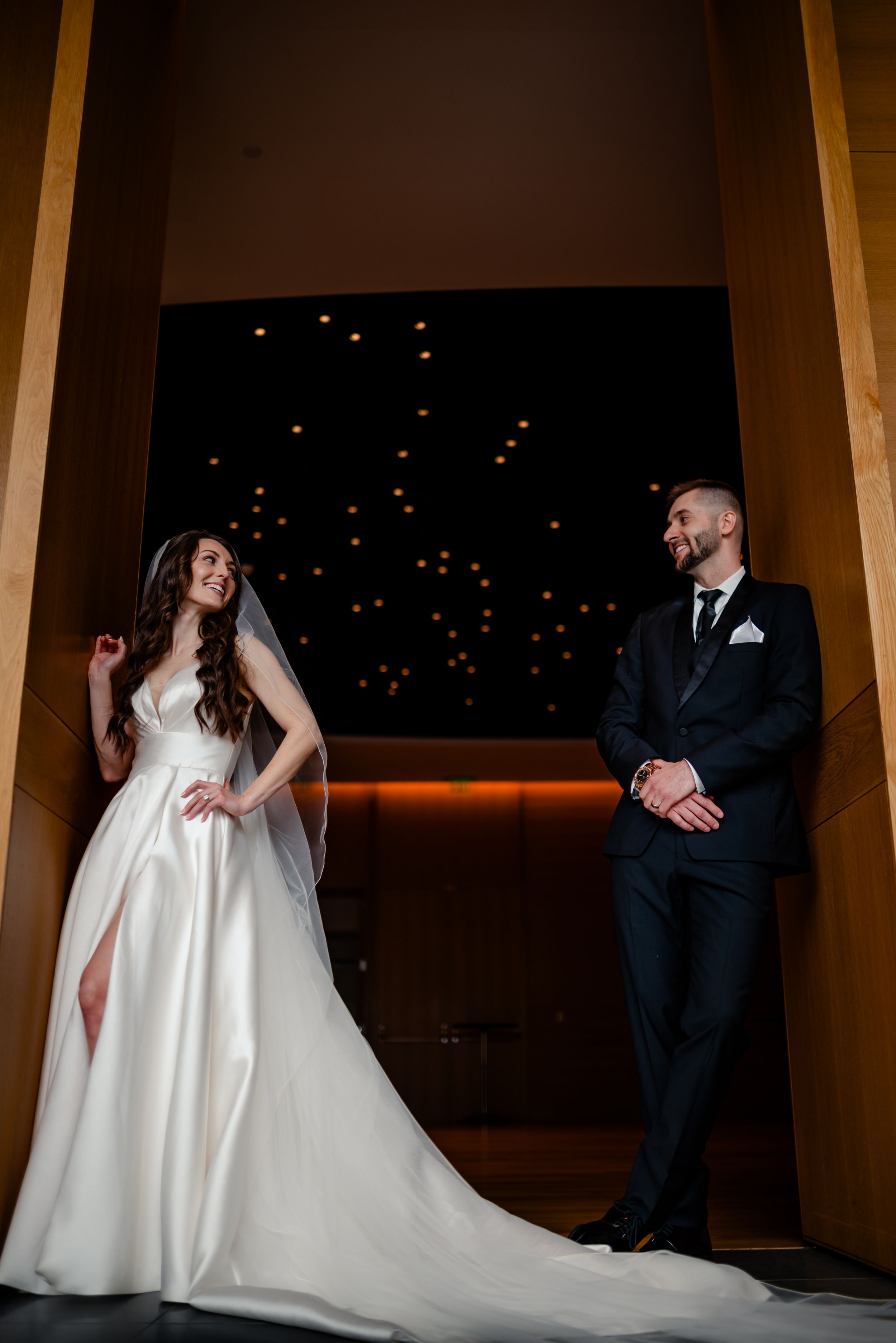 Bride and groom in elegant attire stand apart smiling in dramatic dimly lit wedding venue.