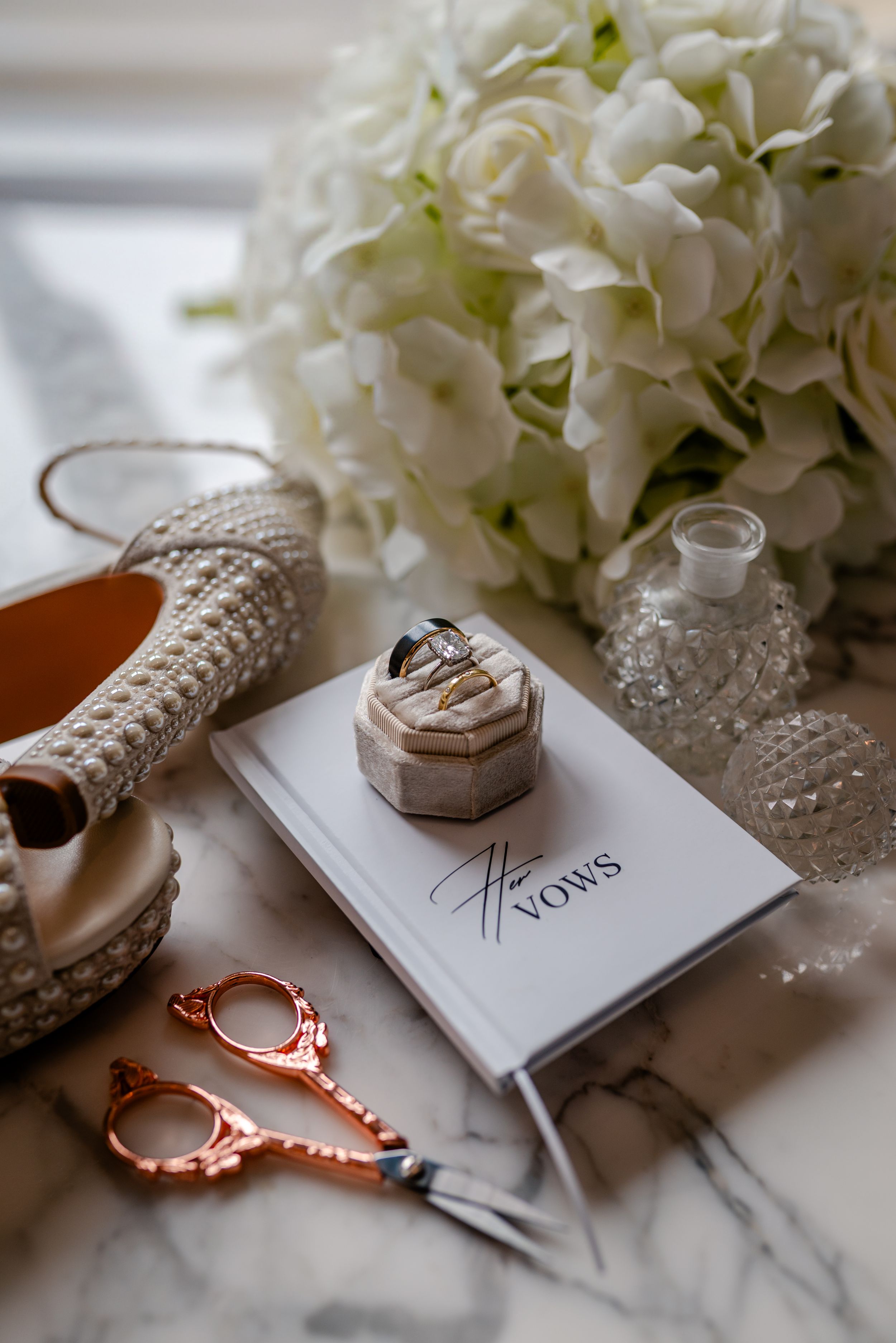 Wedding flat lay with vows book, rings, pearl heels, scissors, and white hydrangea bouquet on marble.