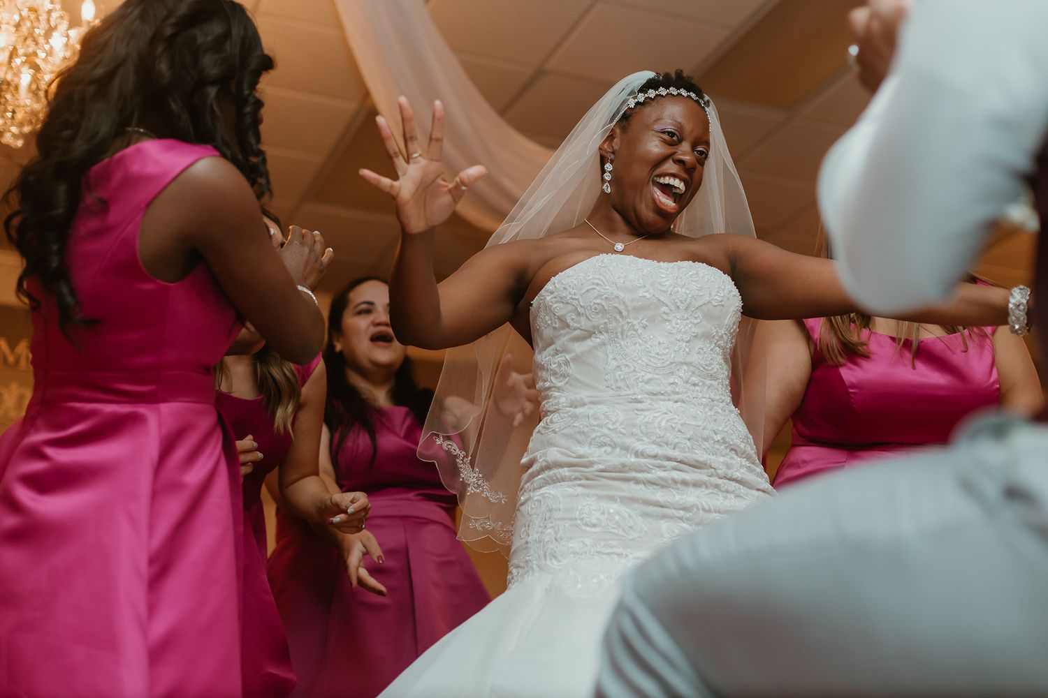 Joyful bride in white lace gown and veil dancing with bridesmaids in pink satin dresses at wedding reception.