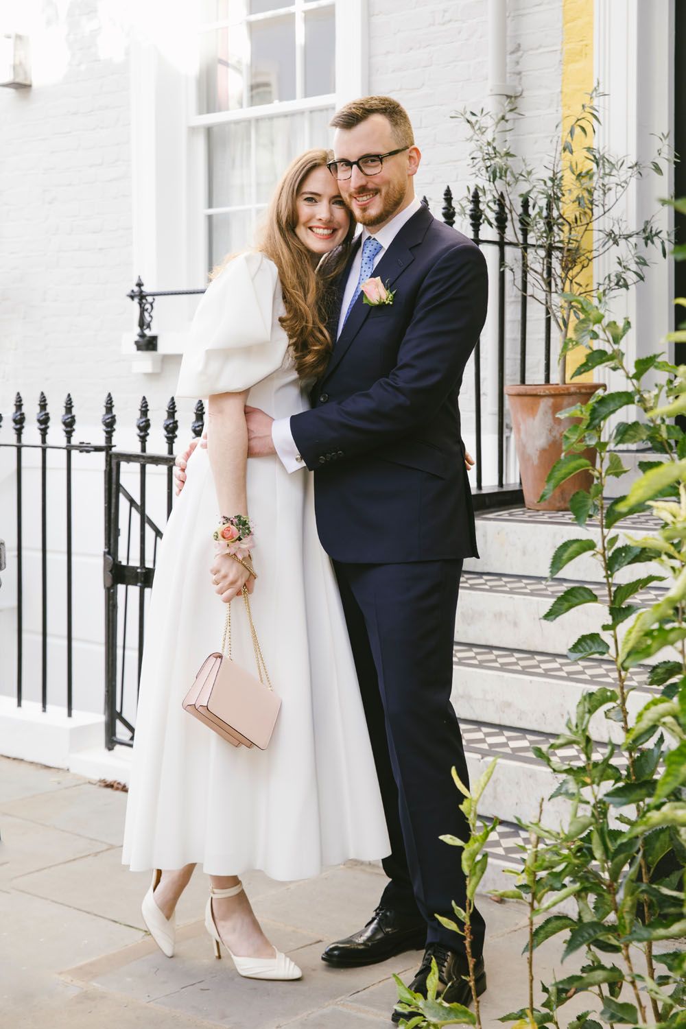 Bride in white midi dress and groom in navy suit embracing outside a white townhouse on their wedding day.