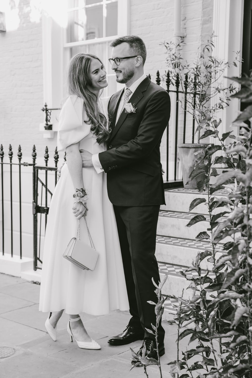 Black and white photo of a smiling bride and groom embracing outside an elegant building on their wedding day.