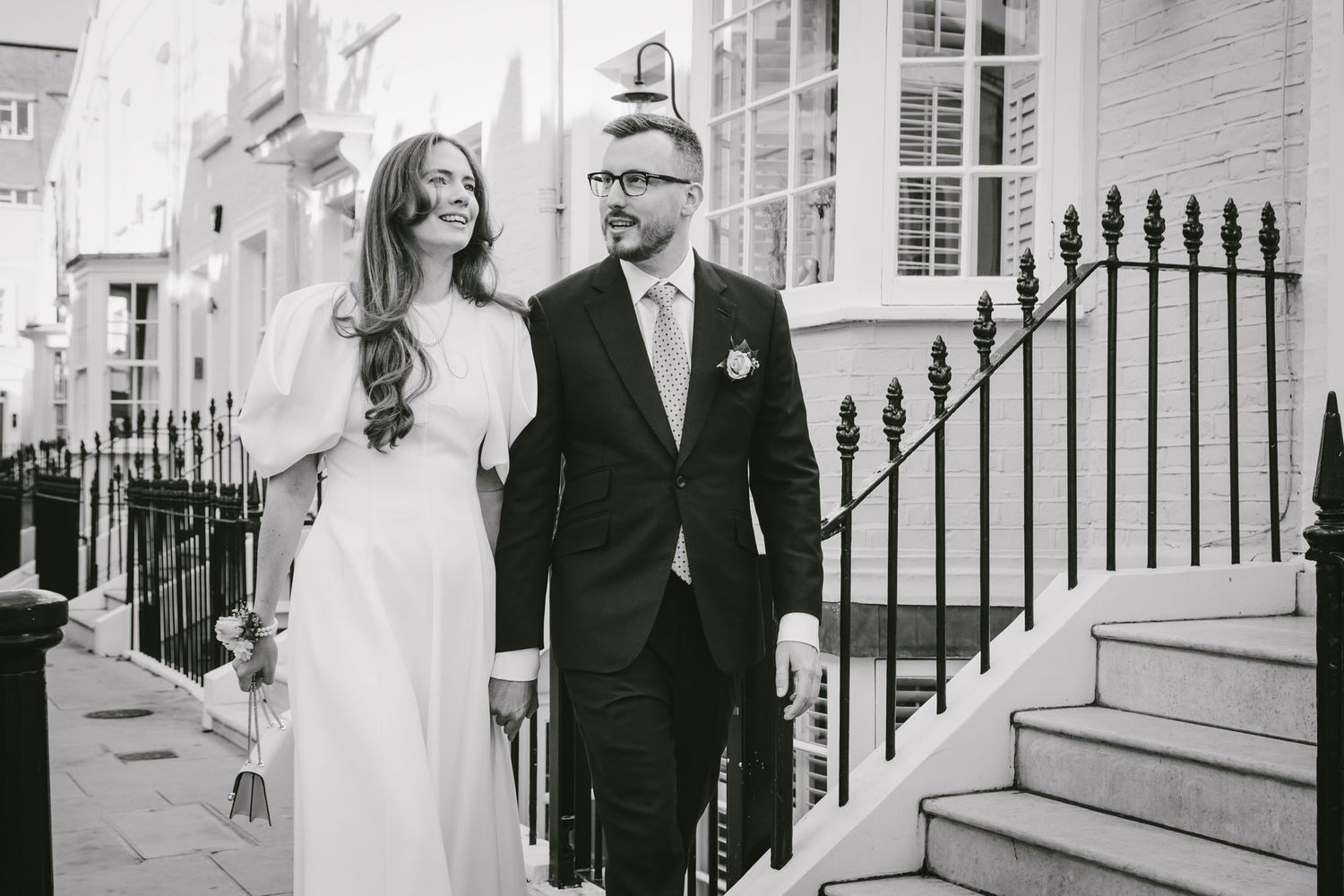 Black and white photo of a bride and groom walking together on a elegant townhouse street on their wedding day.