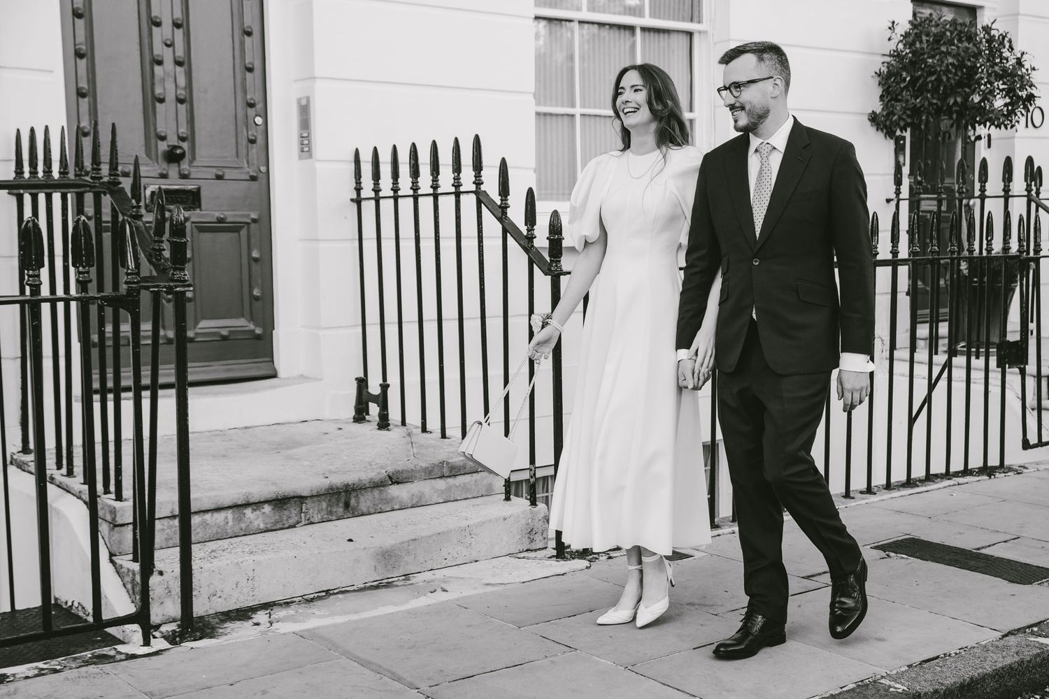 Black and white photo of a couple holding hands walking past iron railings and a Georgian townhouse.