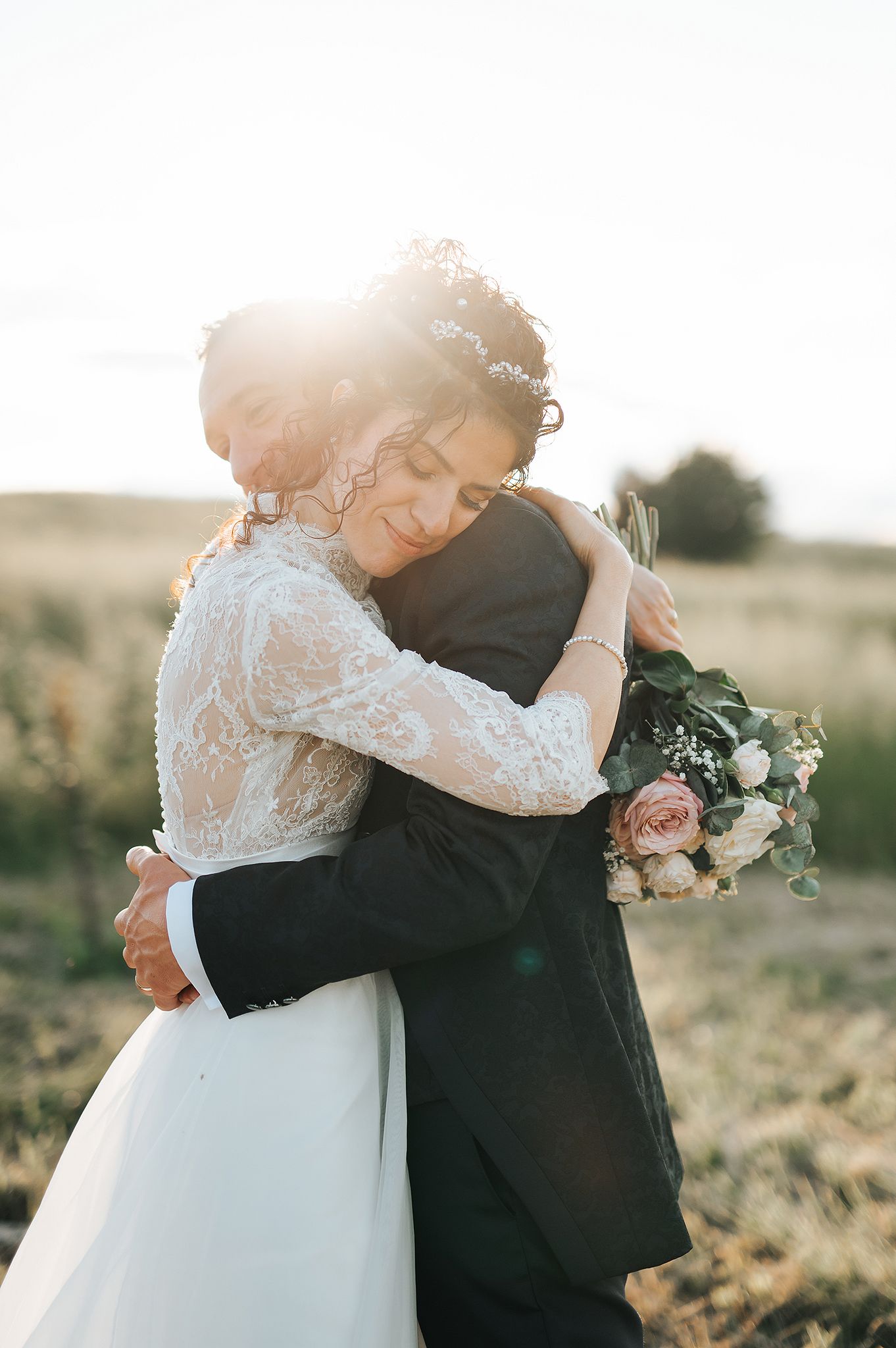 Bride and groom sharing a warm embrace in golden sunset light in an open field.