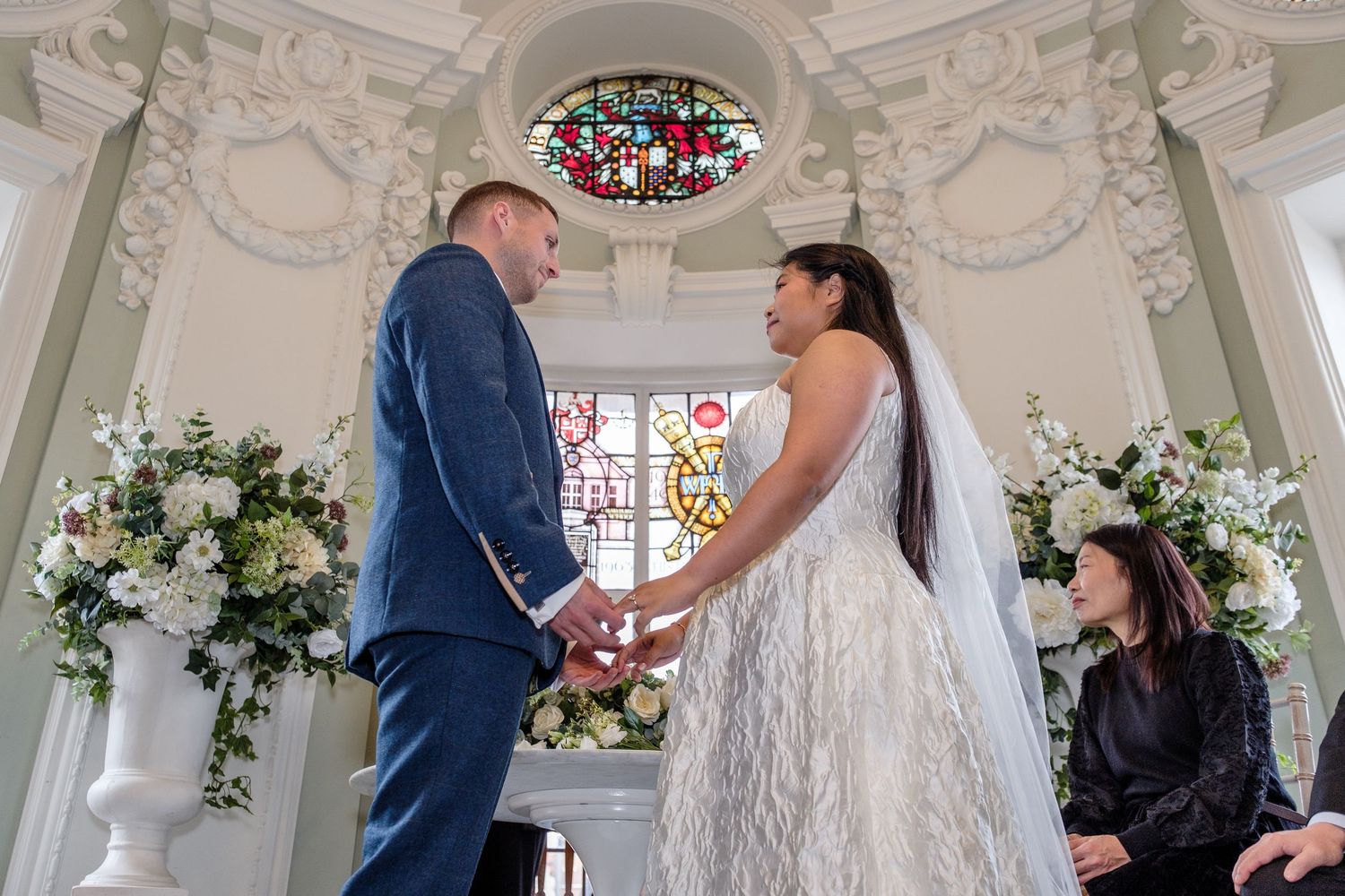 Bride and groom exchange vows holding hands at elegant chapel altar with white floral arrangements and stained glass window.