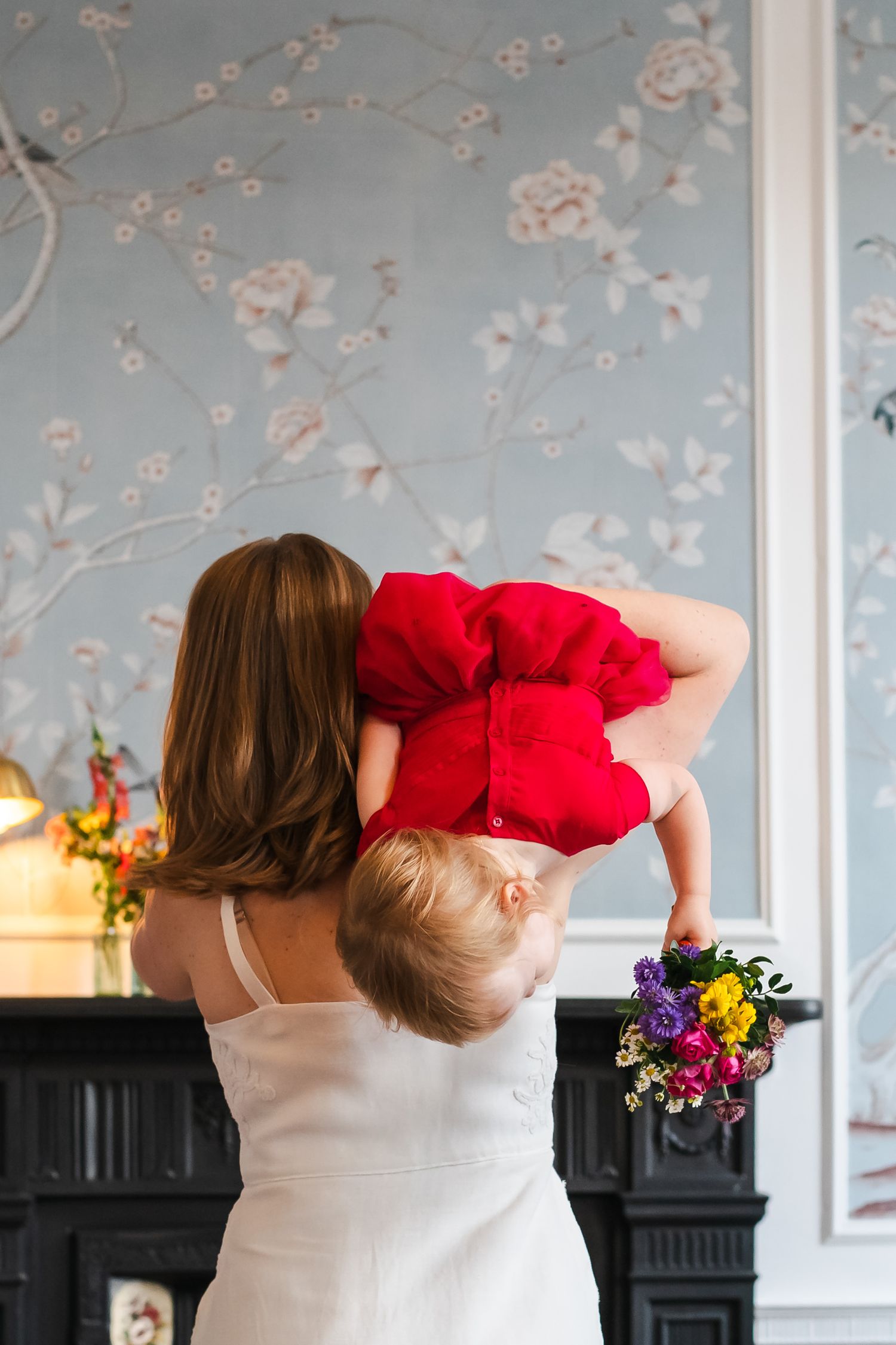 A mother in a white dress holds her baby in a red outfit upside down in an elegant floral wallpapered room.