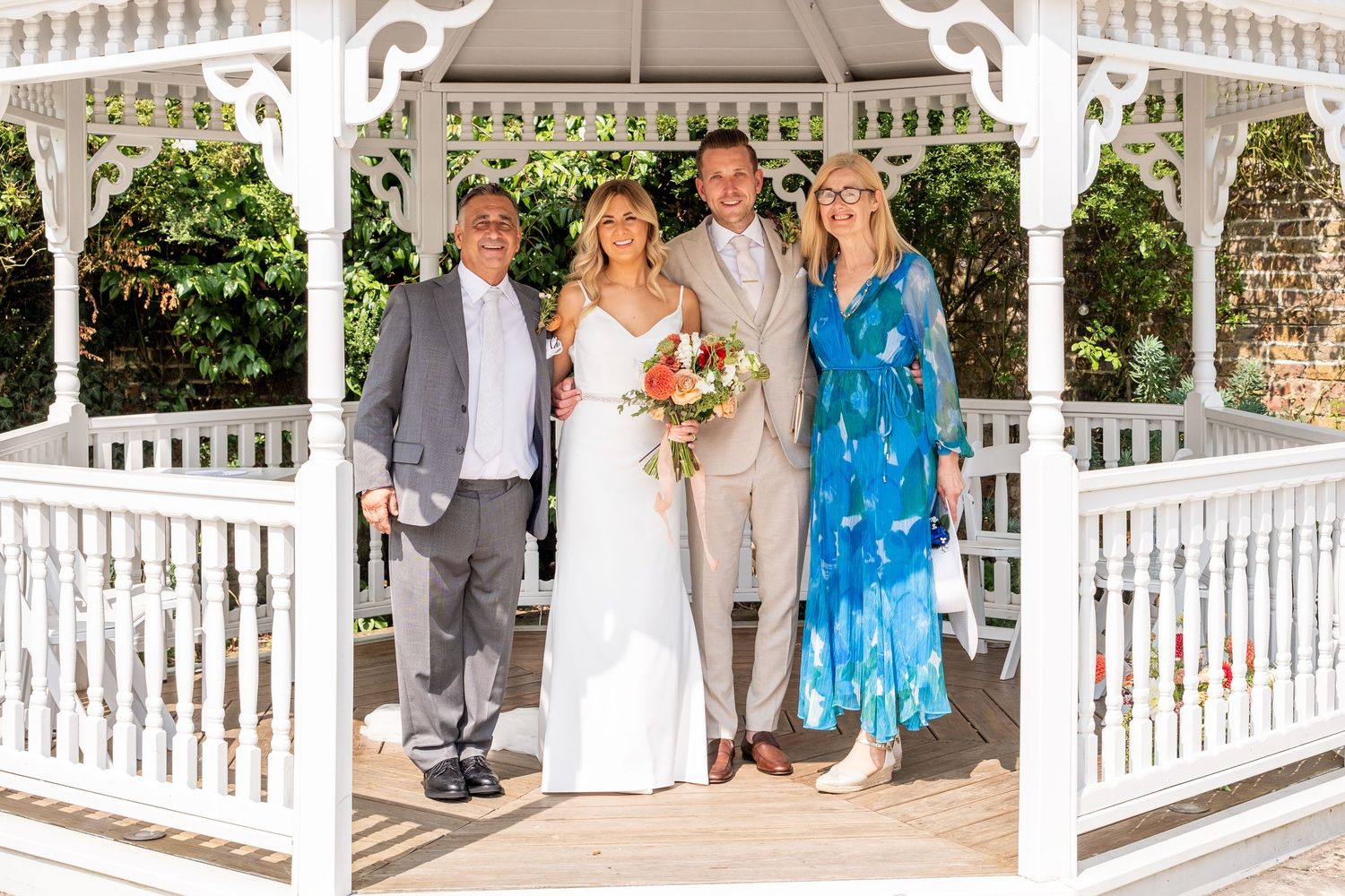 Wedding party of four posing under white gazebo, bride in white gown, groom in tan suit, outdoors.
