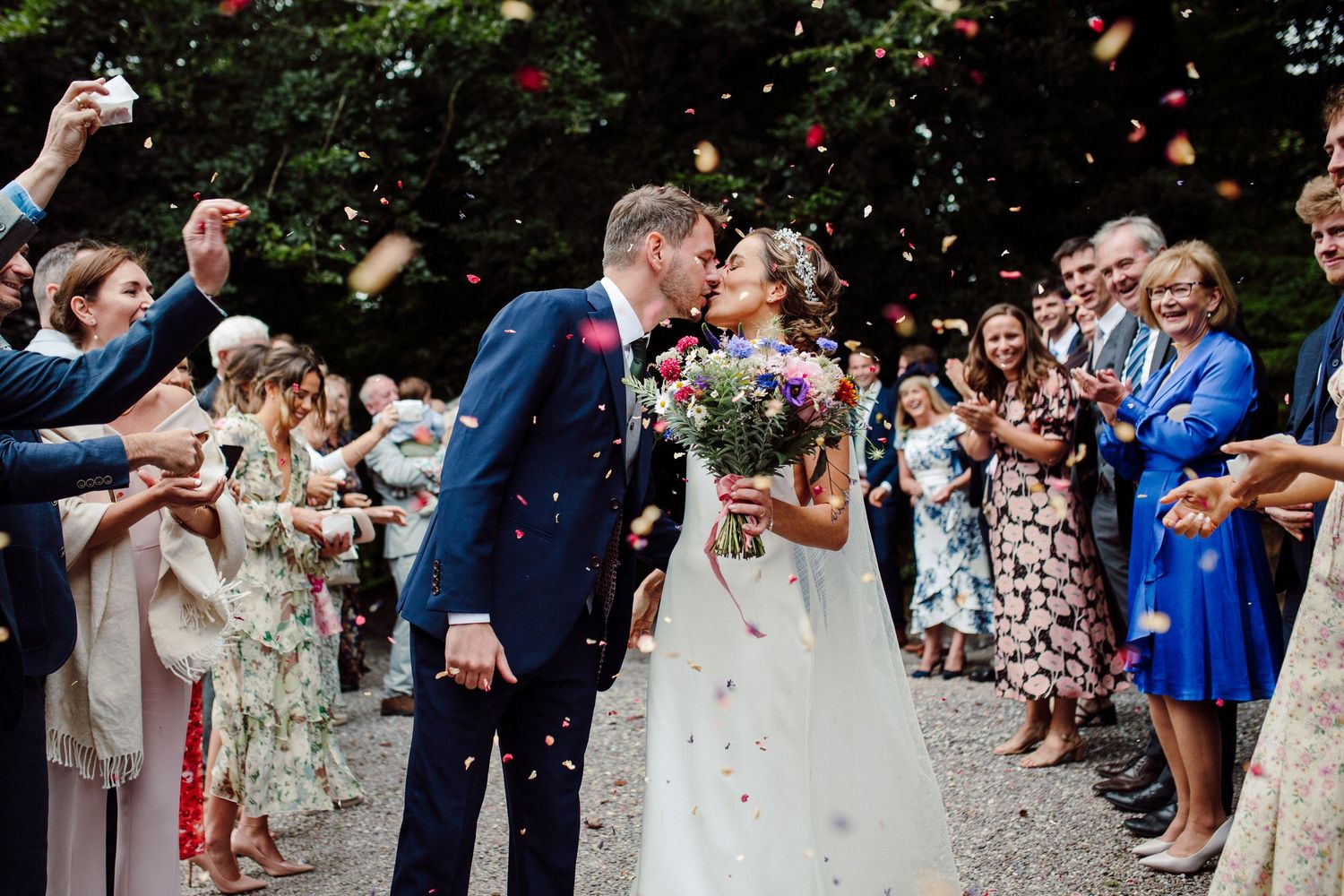 Bride and groom share a kiss while guests throw colorful confetti during a joyful outdoor wedding ceremony.