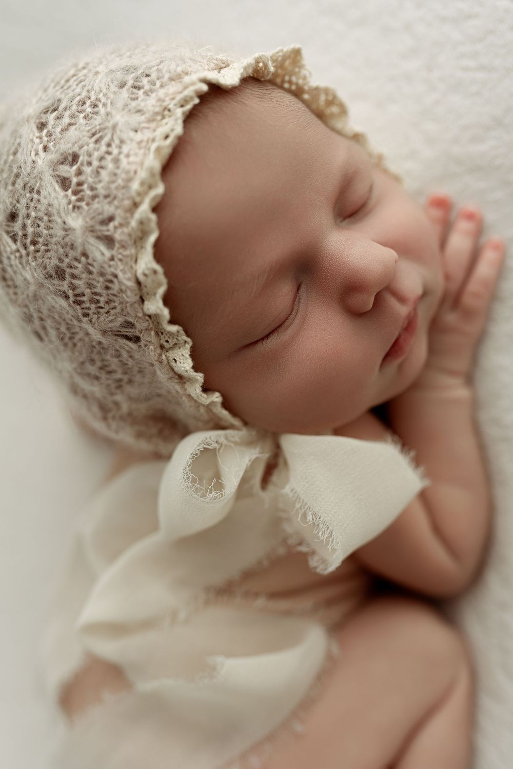 Sleeping newborn baby wearing a delicate cream lace bonnet with ribbon tie, peacefully resting on white fabric.