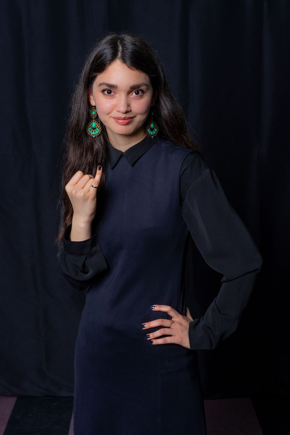 Young woman in navy dress with green earrings posing confidently against dark background.