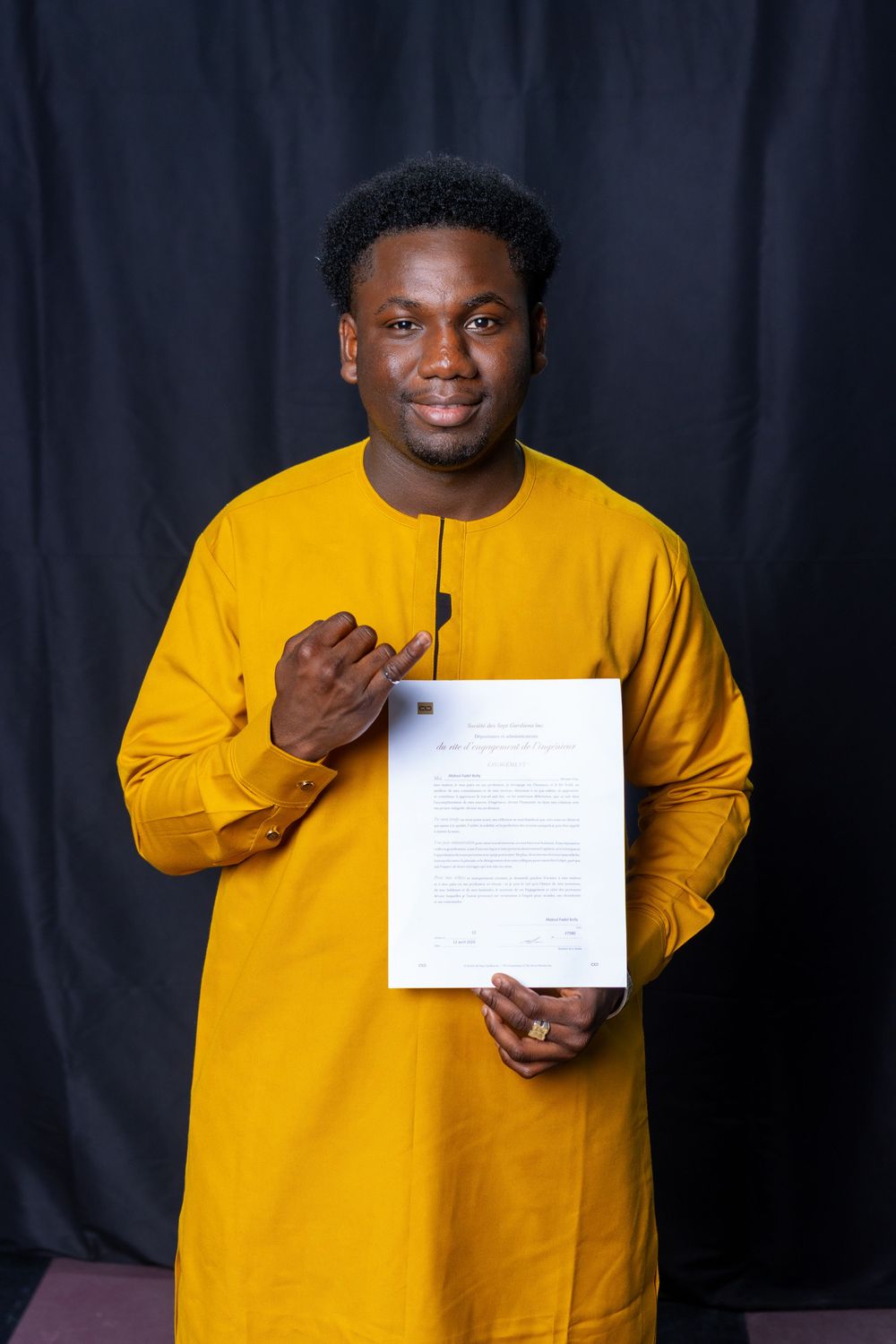 Man in yellow traditional African attire proudly holding and pointing to an official document against dark background.
