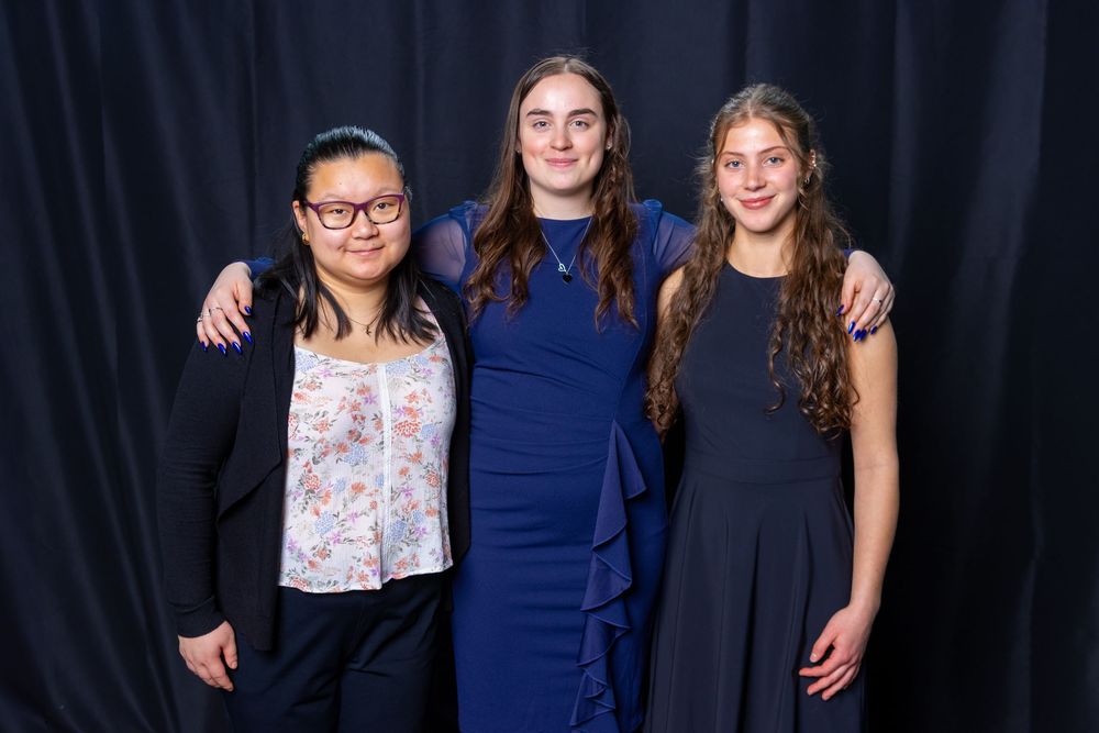 Three young women in formal attire pose together smiling against a dark blue backdrop.