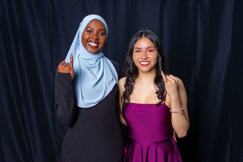 Two smiling women posing together, one wearing a blue hijab and black dress, the other in a purple strapless gown.