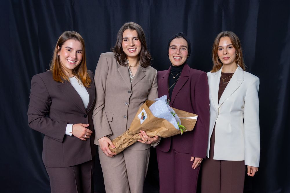 Four professional women in business attire posing together, one holding a bouquet, against a dark backdrop.