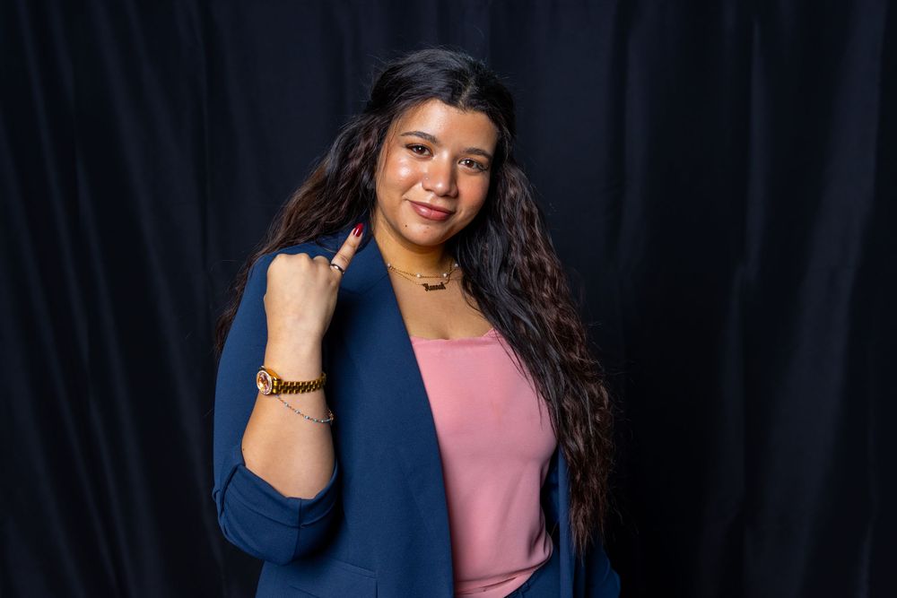 Woman in blue blazer and pink top posing confidently against dark backdrop, wearing gold jewelry.