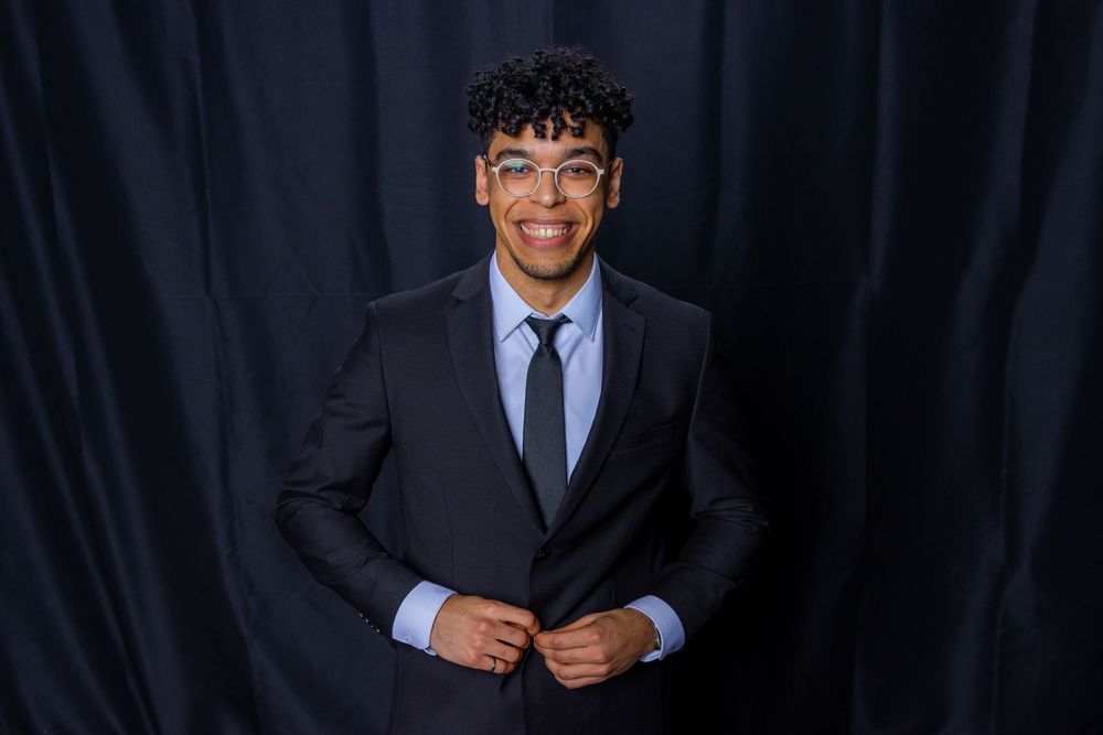 Young man smiling in a black suit and light blue tie, posing against a dark navy backdrop.